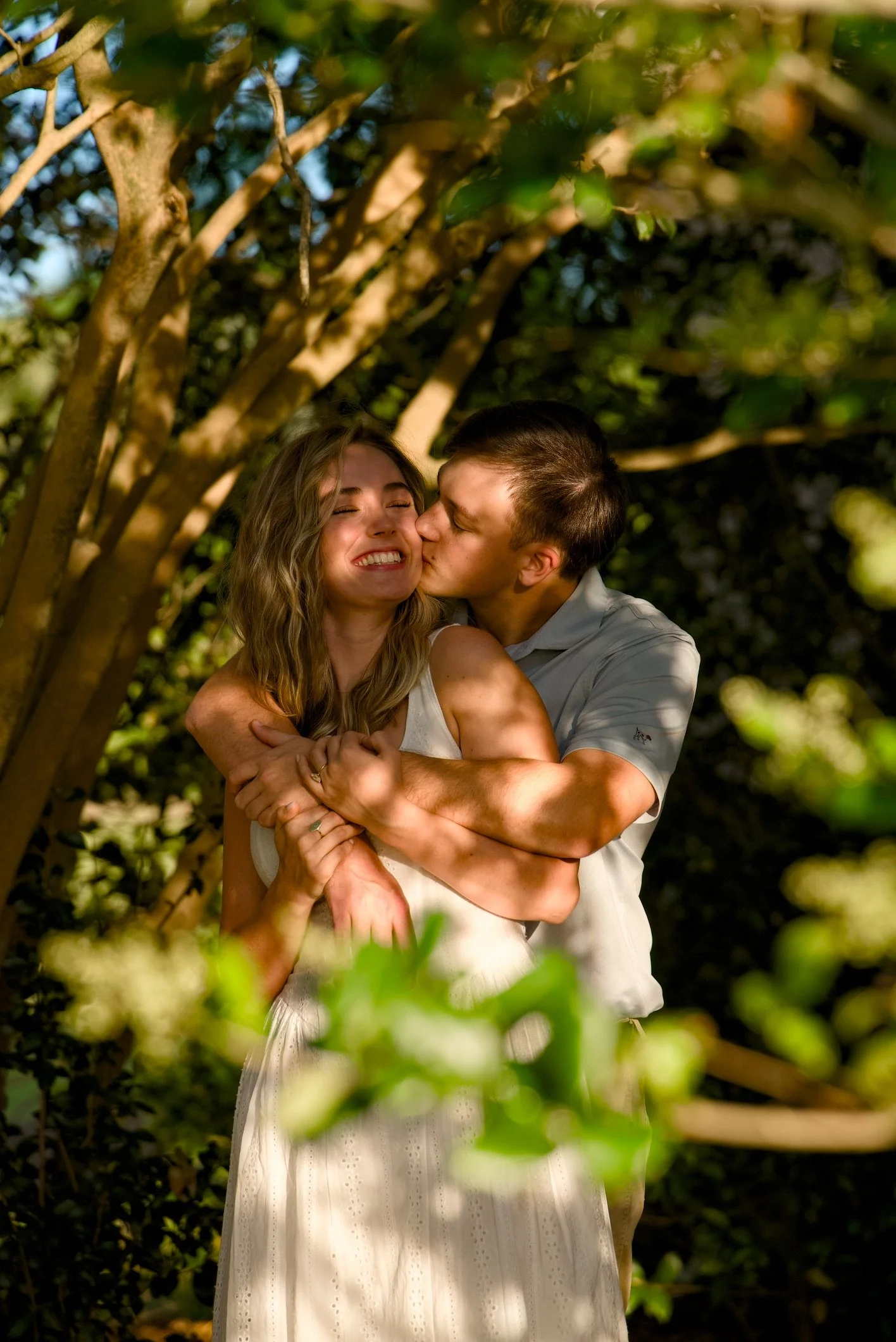 A young man and woman embracing outdoors under tree branches, with sunlight through leaves, the woman smiling with eyes closed, the man kissing her cheek.