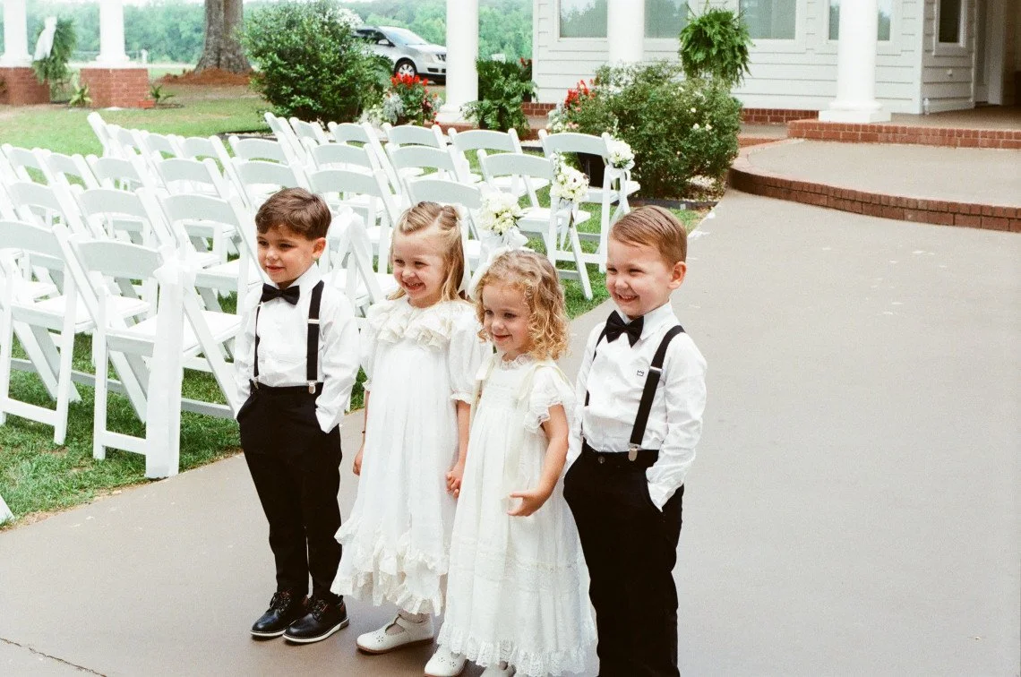 Four children, two boys and two girls, dressed in formal attire, standing in a line outdoors at a wedding or special event, with white chairs and greenery in the background.
