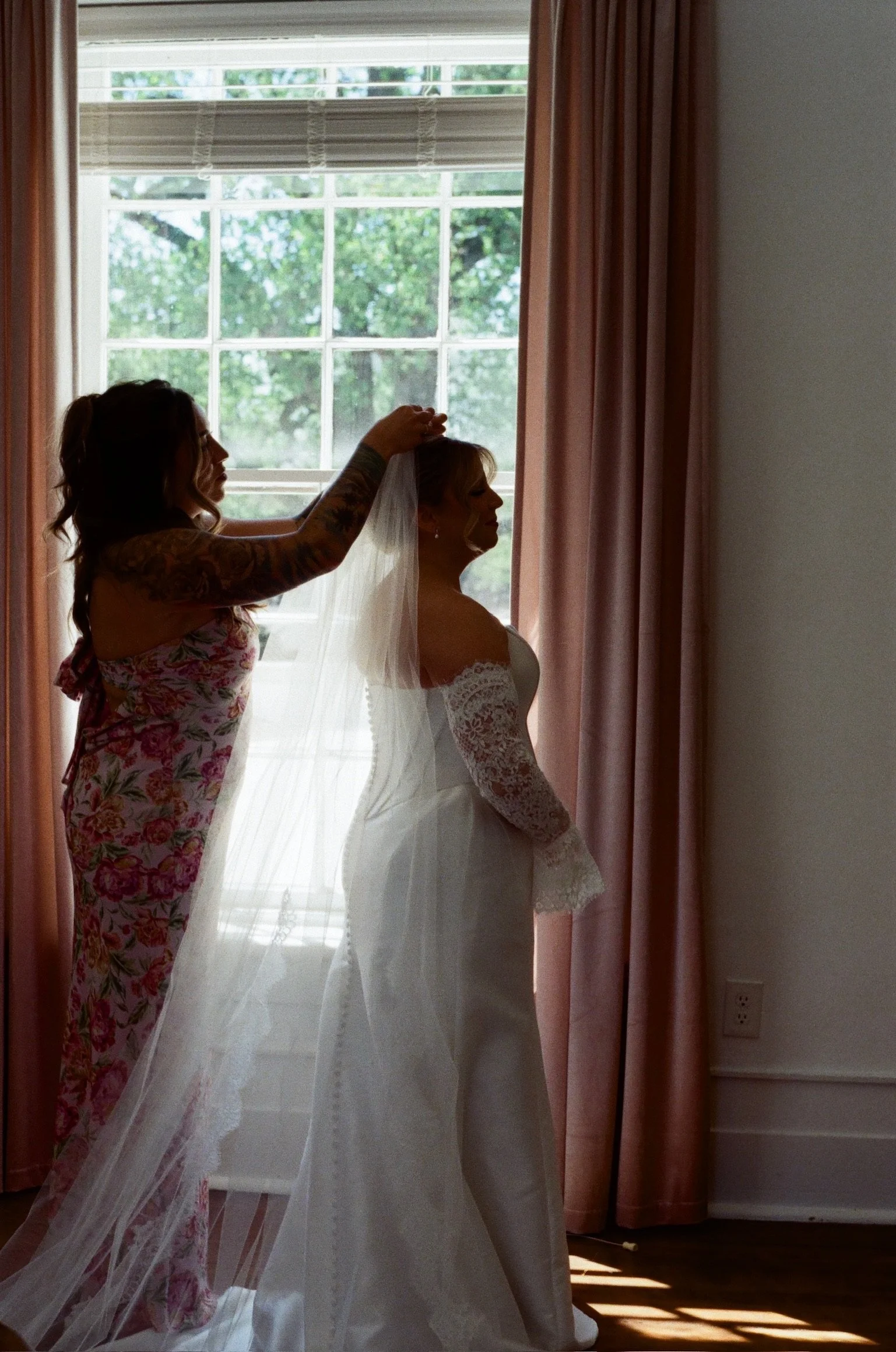 A bride in a white lace dress sitting by a window, with a woman in floral pajamas adjusting her veil, as sunlight filters through pink curtains.
