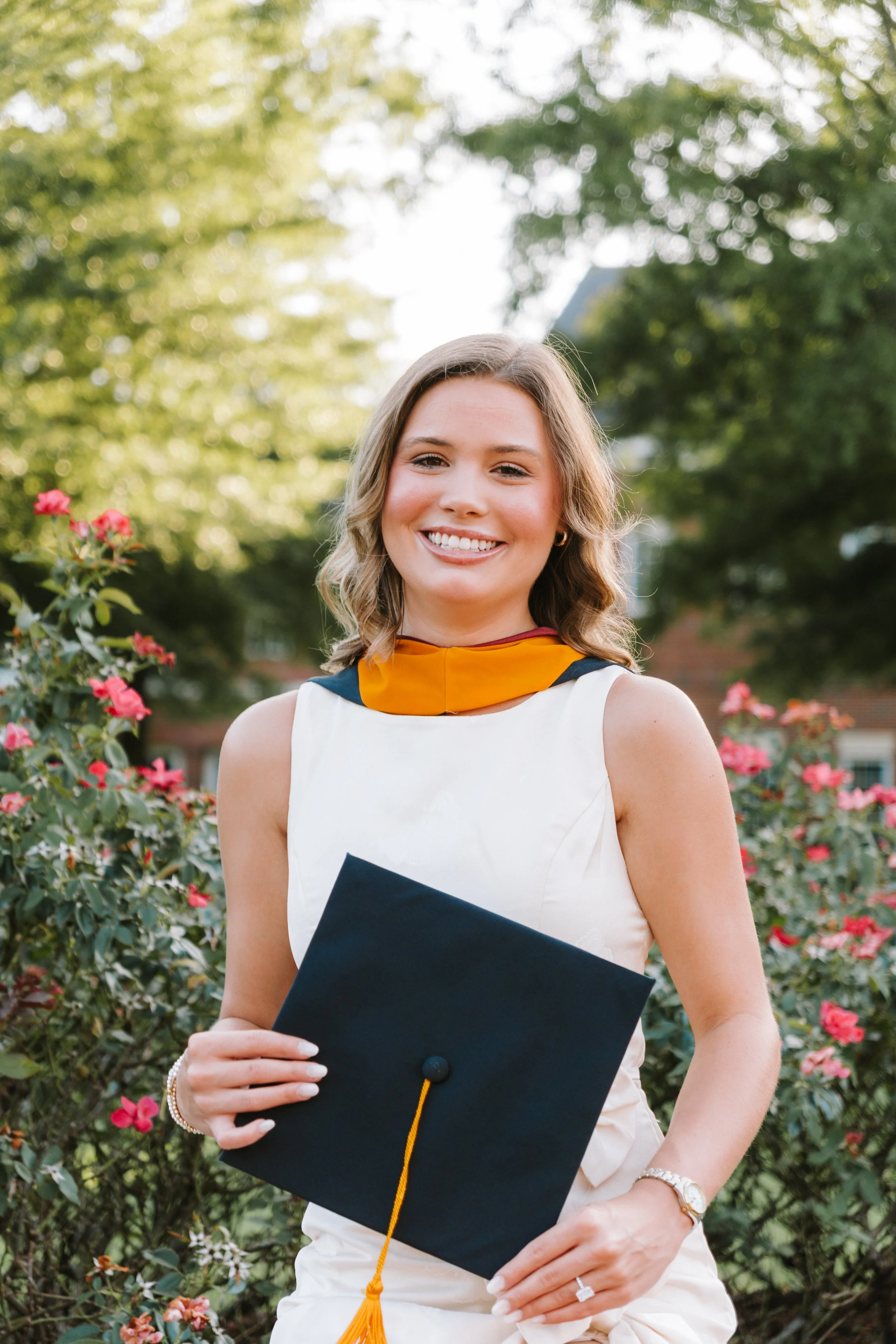 A young woman holding a diploma in a graduation cap, outdoors among pink and red flowers with green trees in the background, smiling at the camera.