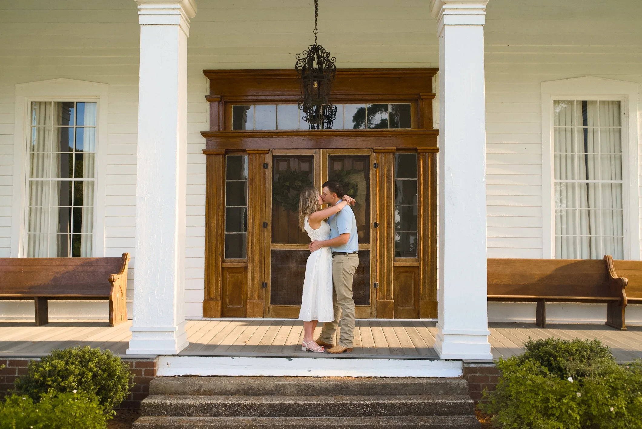 A bride and groom share an intimate moment on the porch of a white house with large windows and wooden doors, surrounded by benches and greenery.