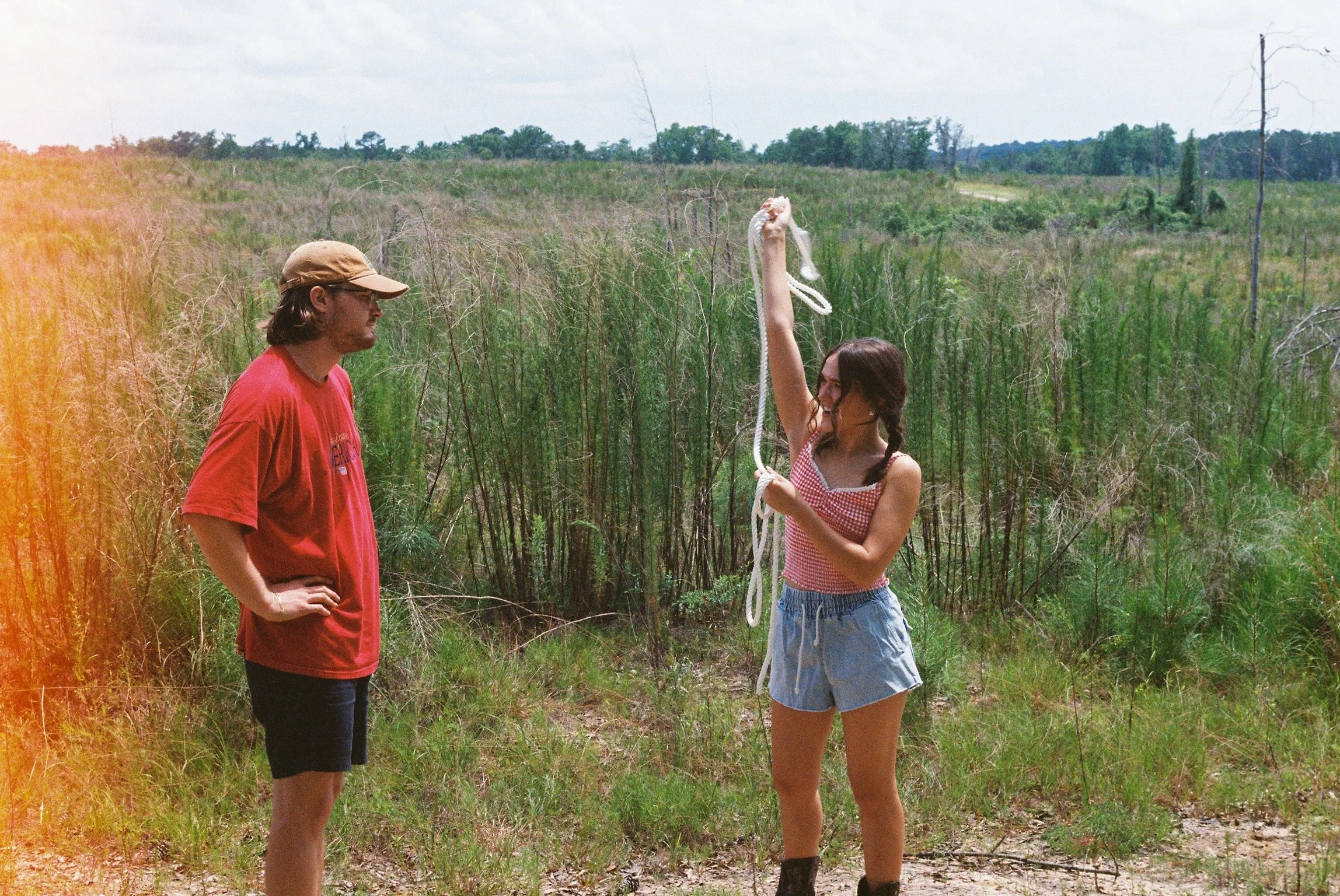 A woman holding a rope with a glass object attached to it, standing in a grassy field with trees in the background, while a man wearing a cap and glasses observes.