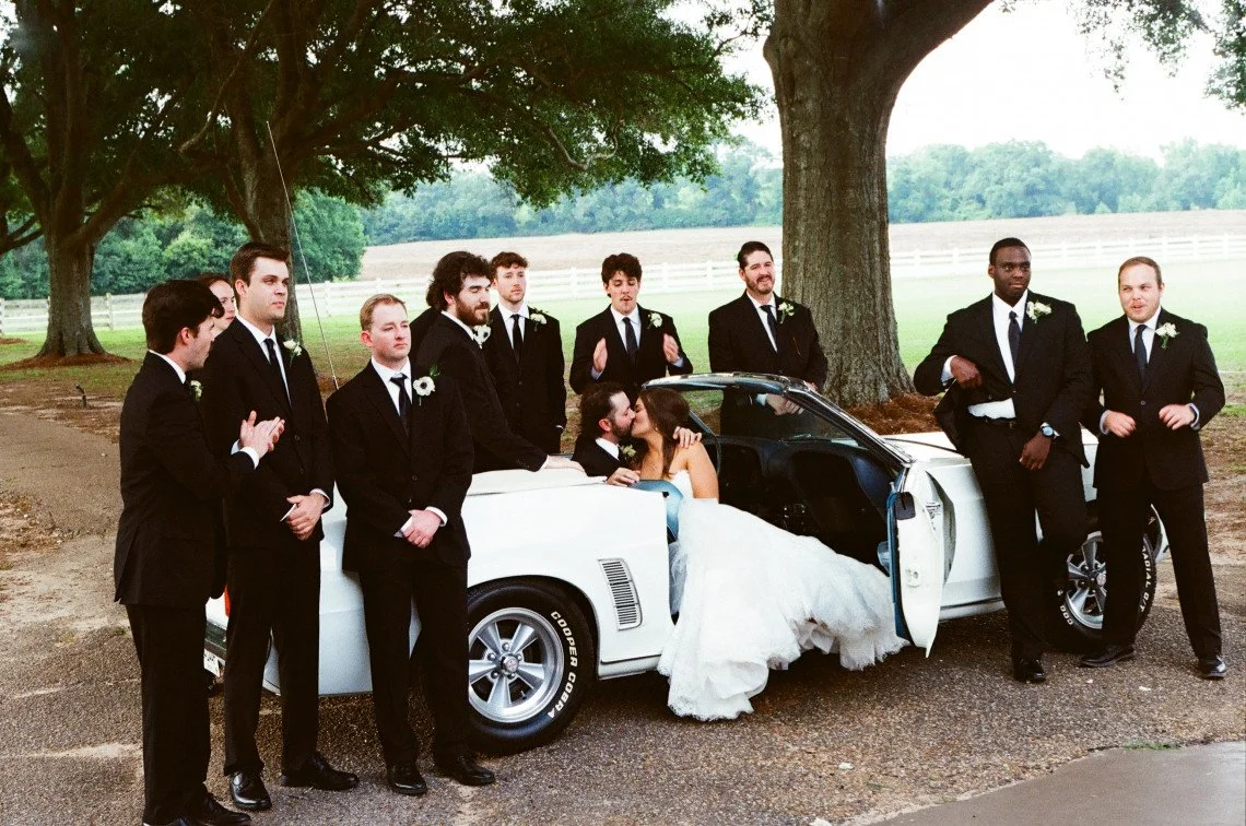 Bride and groom kissing inside a white sports car surrounded by groomsmen in suits outdoors under large trees.