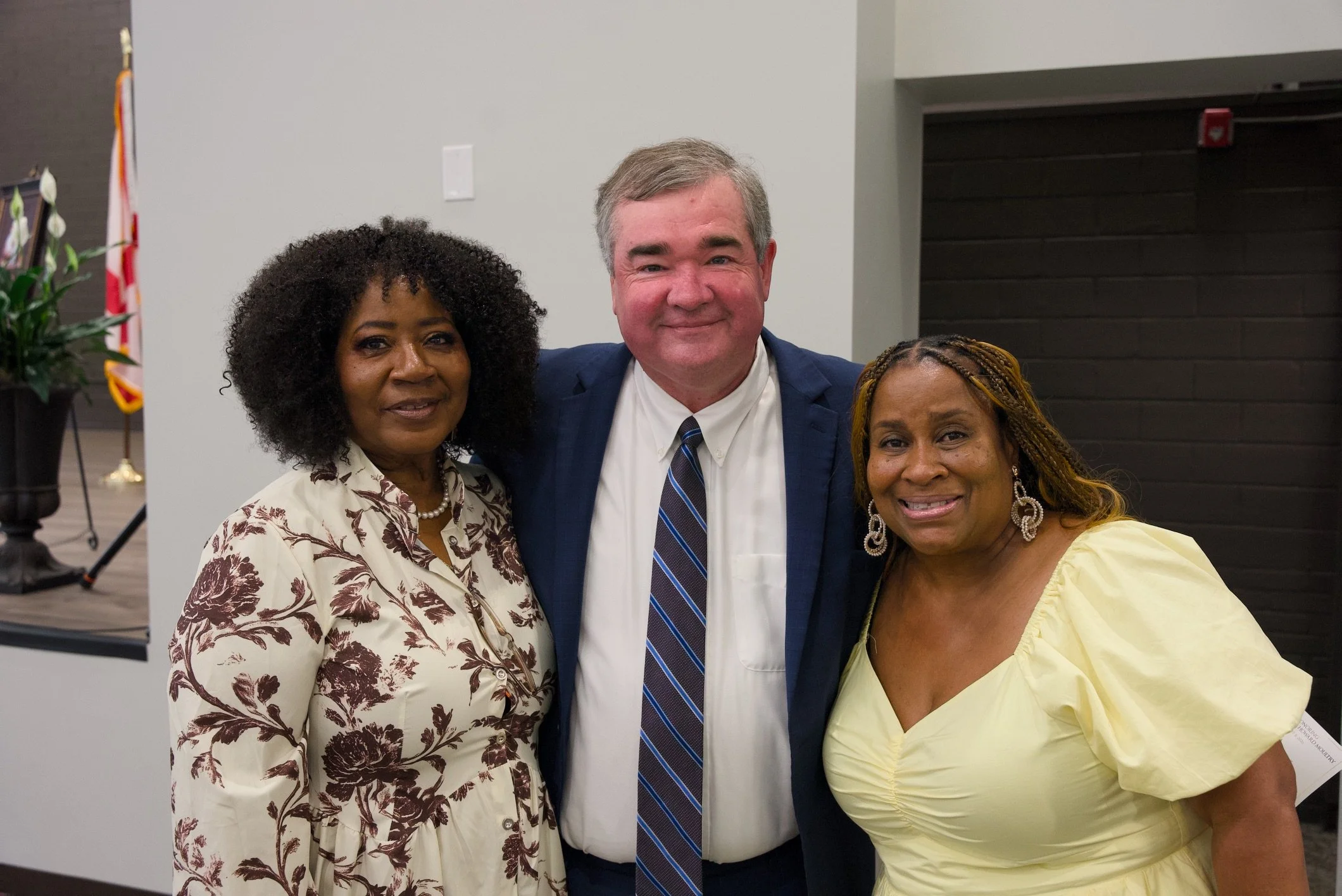Three people standing together indoors, smiling, with an American flag and decorative plants in the background.