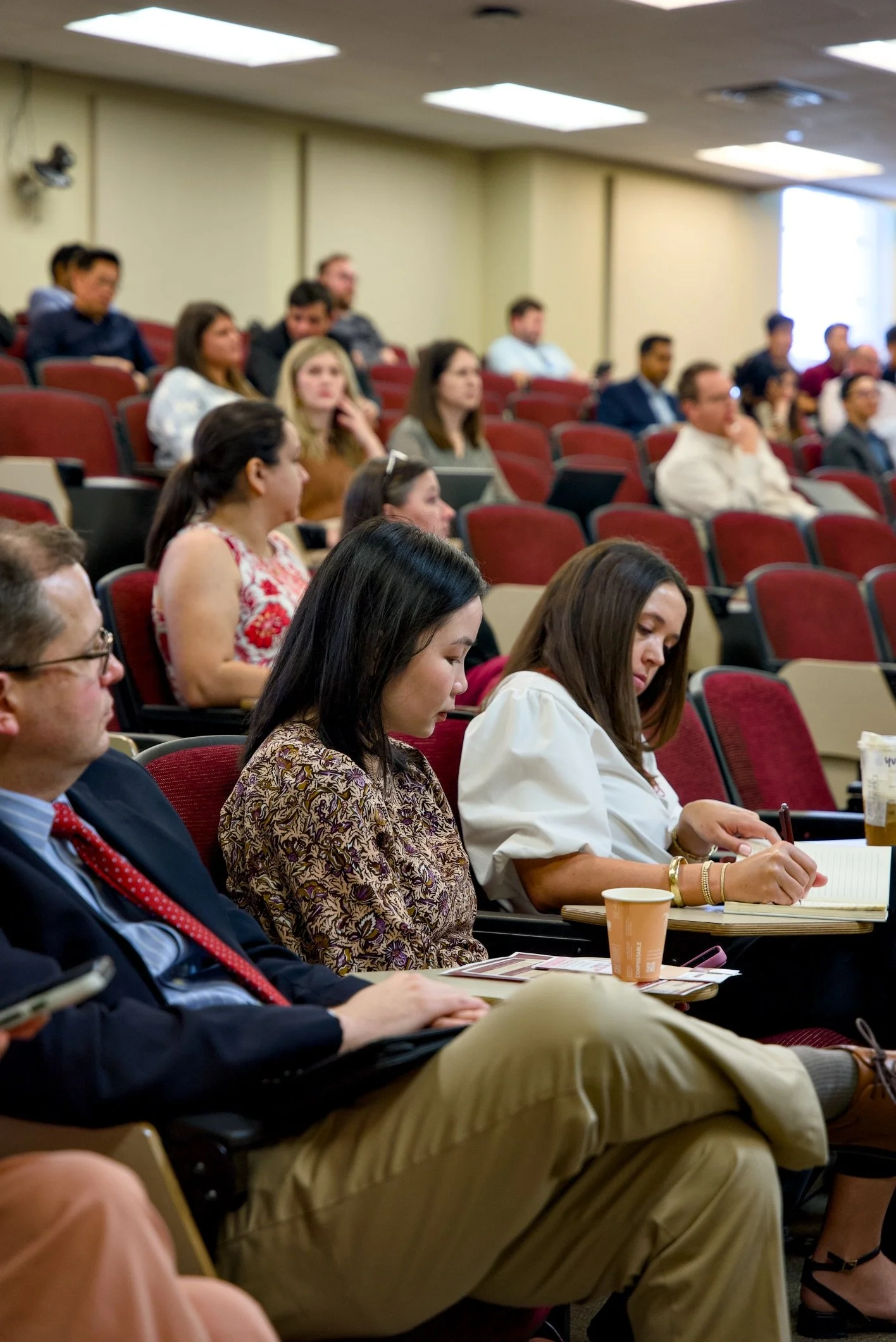 A group of people sitting in a lecture hall, listening to a presentation, with some taking notes and others looking towards the front of the room.