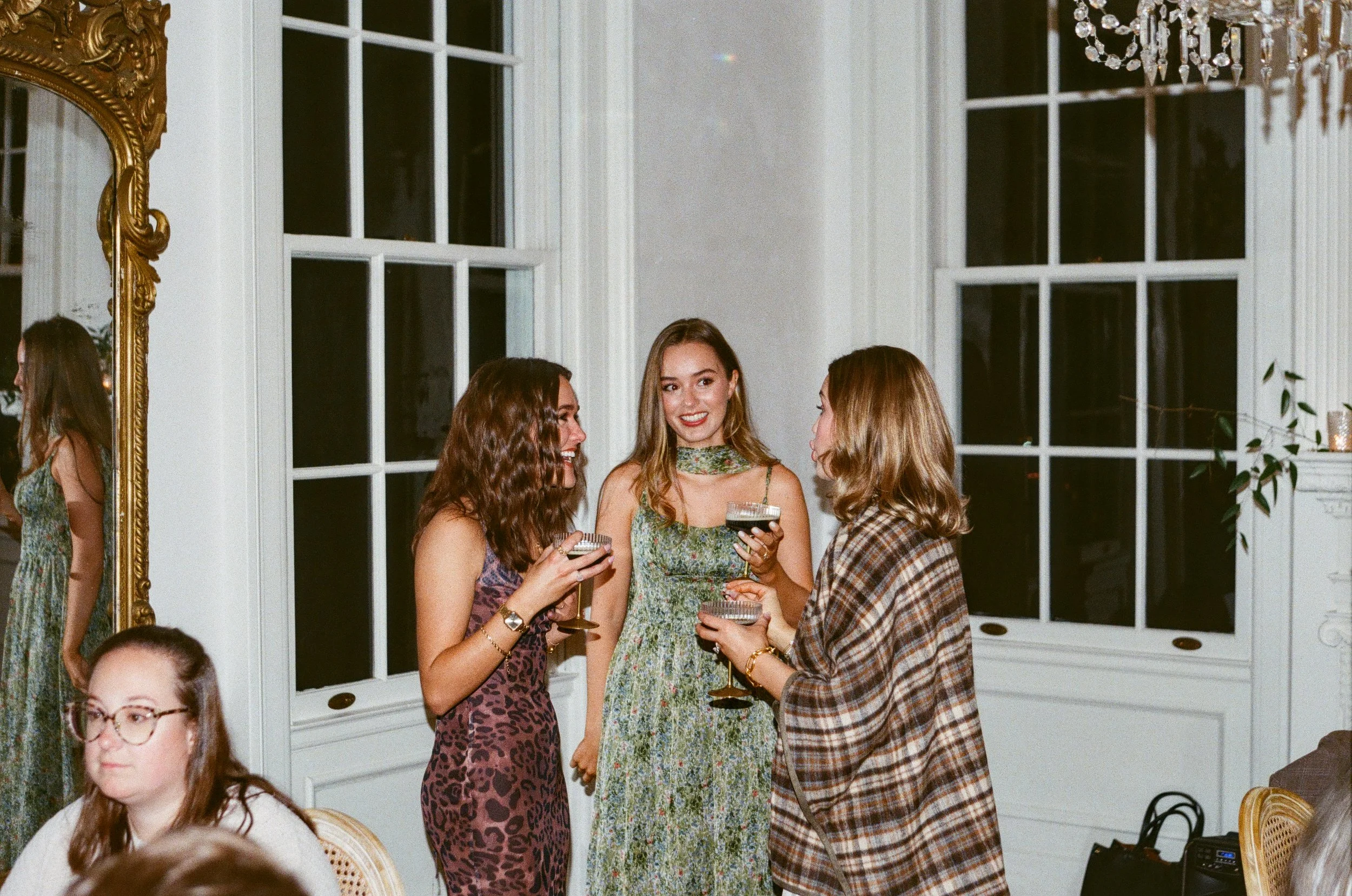 Group of women socializing at an indoor event, holding drinks and talking, in a well-lit room with large windows and a chandelier.