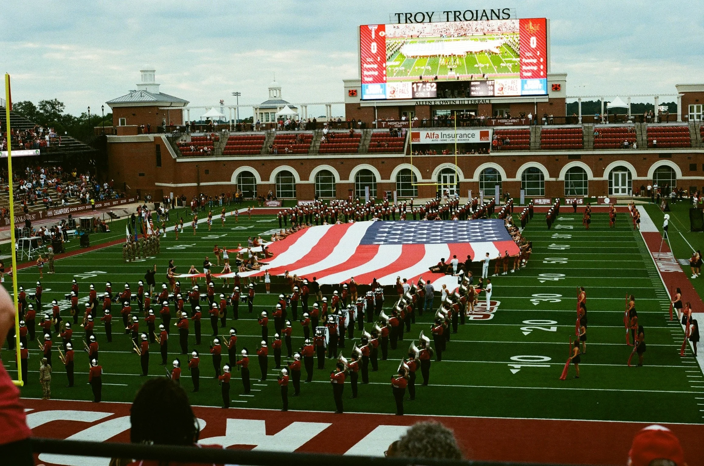 College football stadium with a marching band performing on the field during a game. The stadium has spectators in the stands and a large scoreboard displaying the game score and time remaining. The field is decorated with the American flag.
