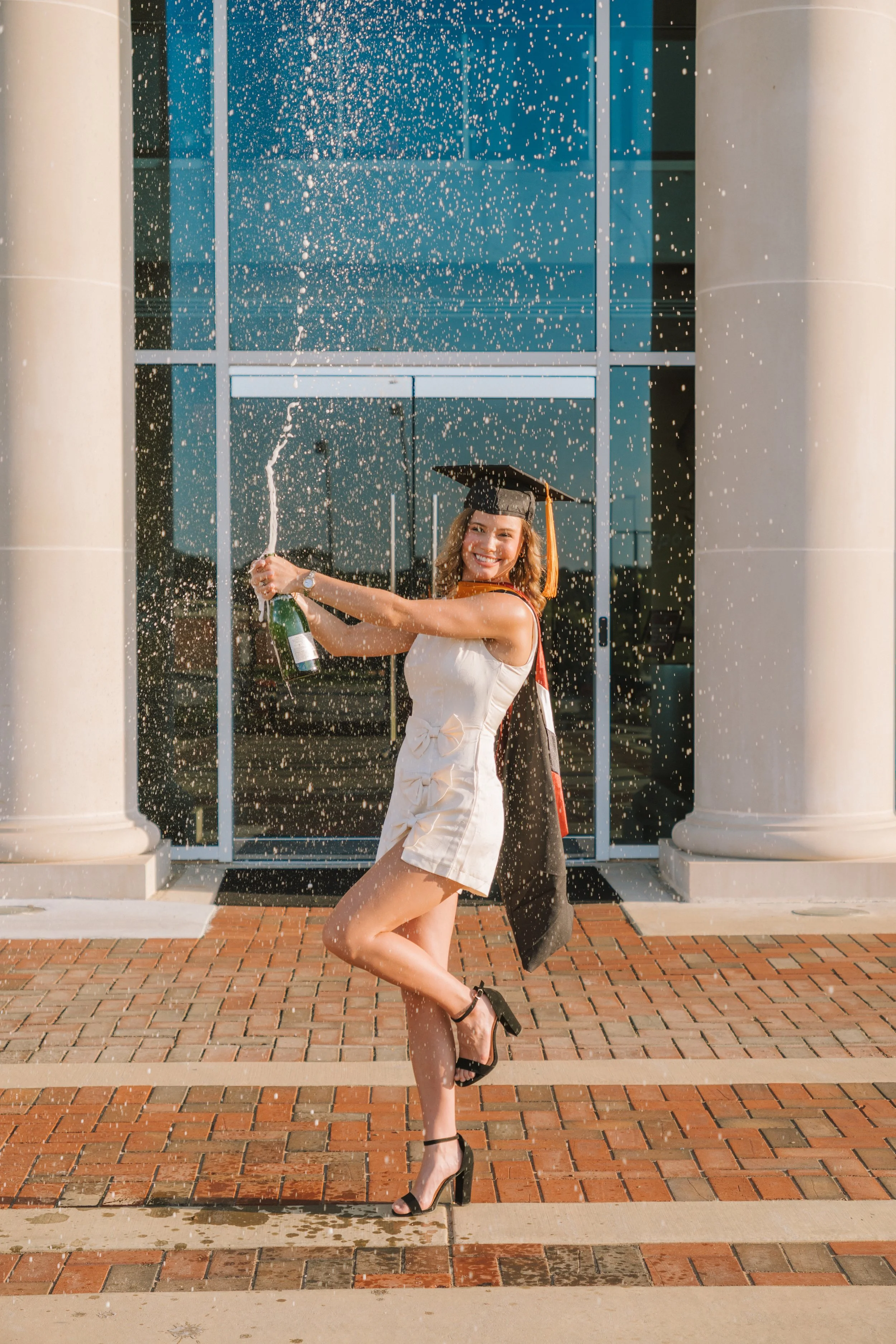 A joyful woman in a white dress and black heels celebrating graduation outdoors, spraying champagne and standing on a brick sidewalk in front of a building with large glass windows and columns, with celebratory sparks flying around.