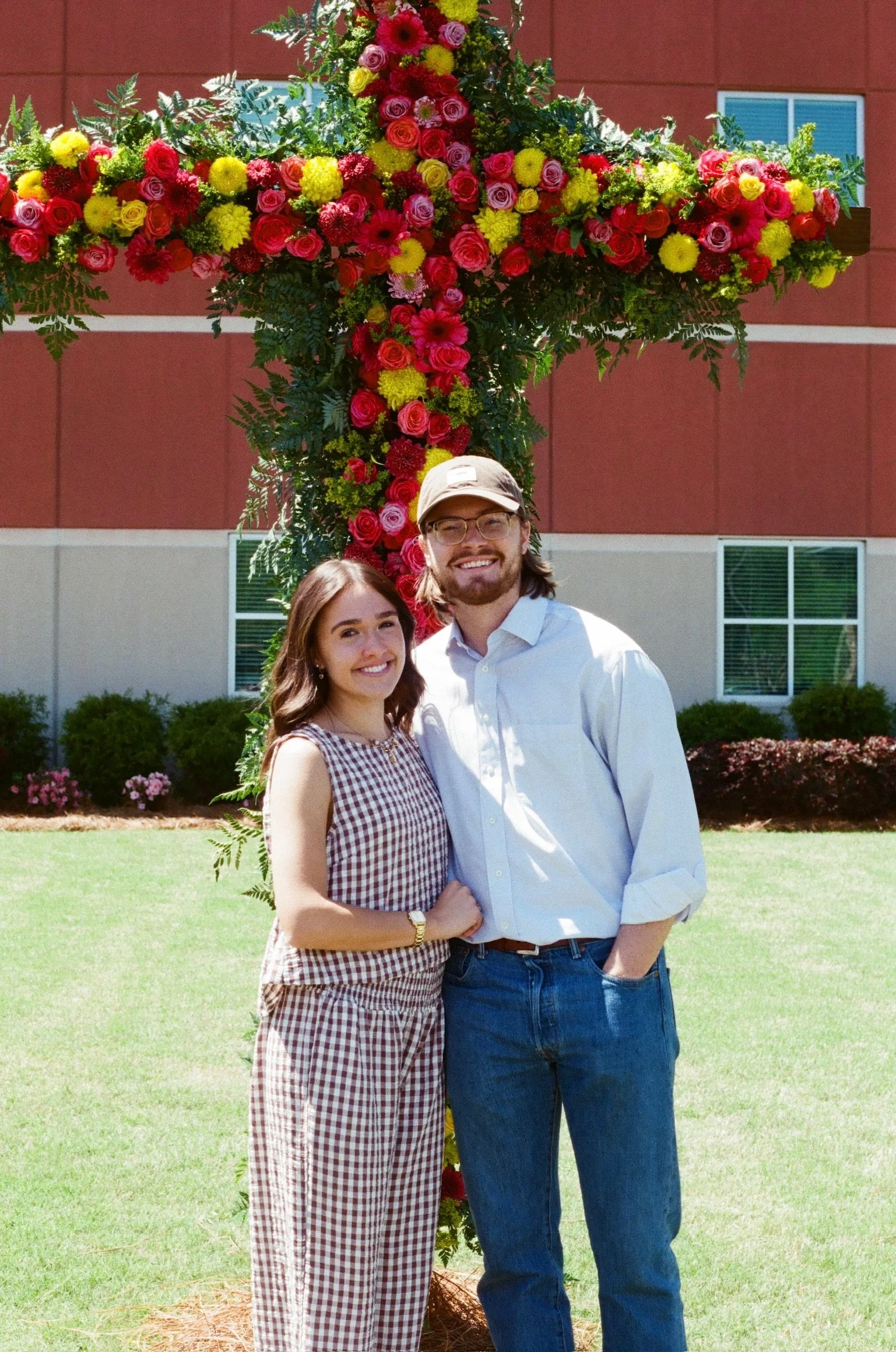 A smiling couple standing outdoors in front of a large floral cross decoration with pink, red, and yellow flowers, on a lawn in front of a building with red siding and white windows.