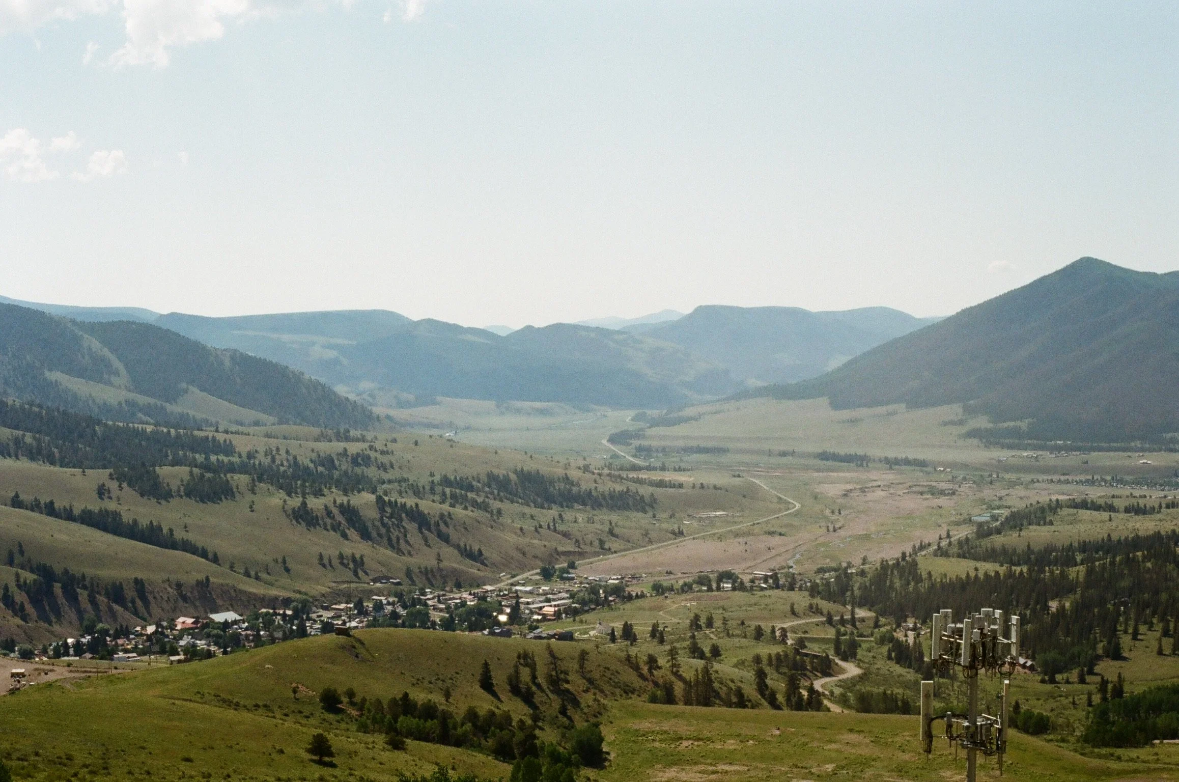 A panoramic view of rolling green hills and mountains under a partly cloudy sky, with a small town in the valley and electrical poles in the foreground.
