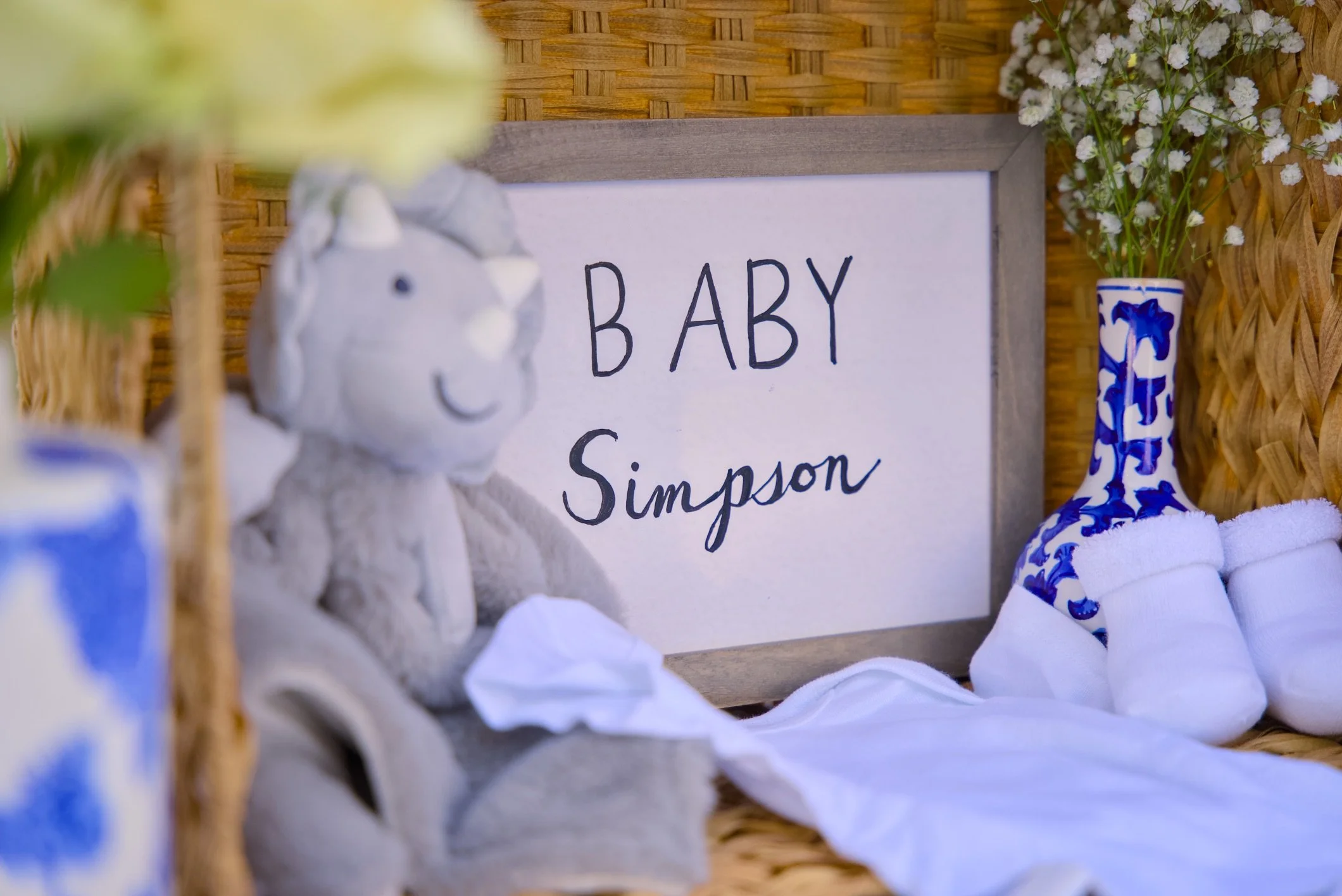 A small stuffed lamb toy, a framed sign reading "BABY Simpson," a blue and white patterned vase with white flowers, and blue and white decorative items in a wicker basket setting.