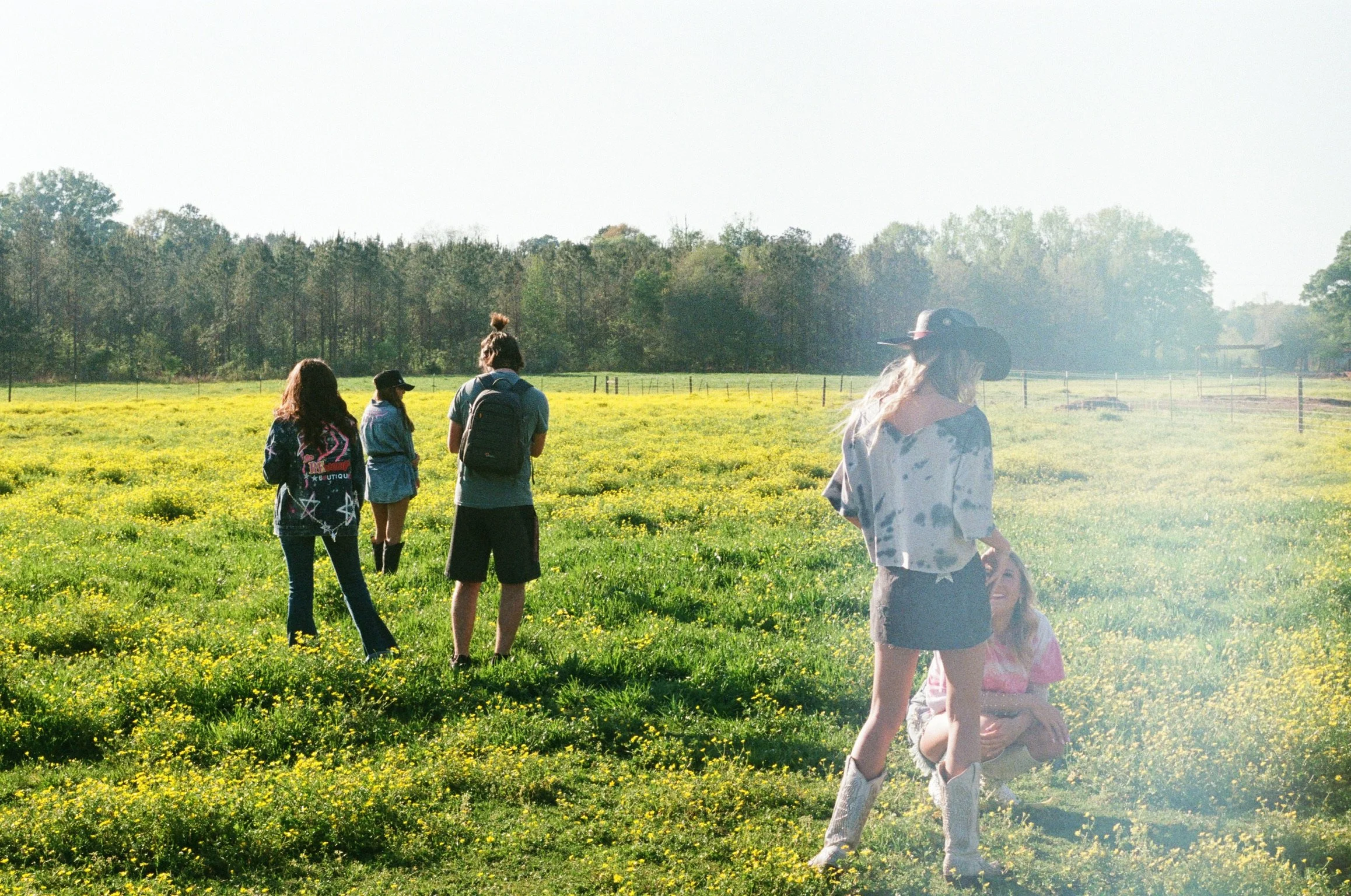 Group of young women standing and sitting in a grassy field with yellow flowers, near a tree line, enjoying sunny weather.