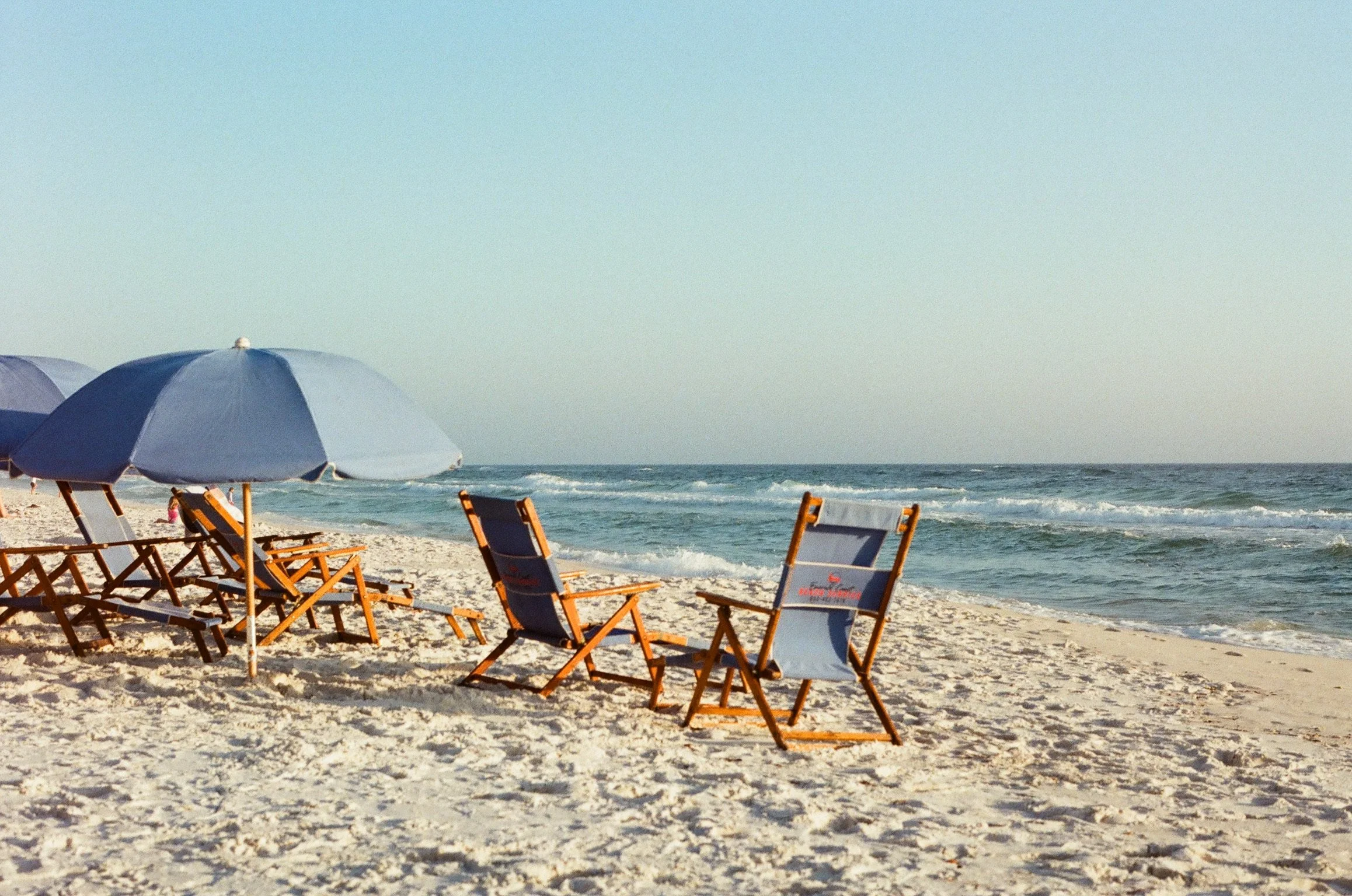 Beach with blue umbrellas and orange lounge chairs on sandy shore, ocean waves in background under clear sky.