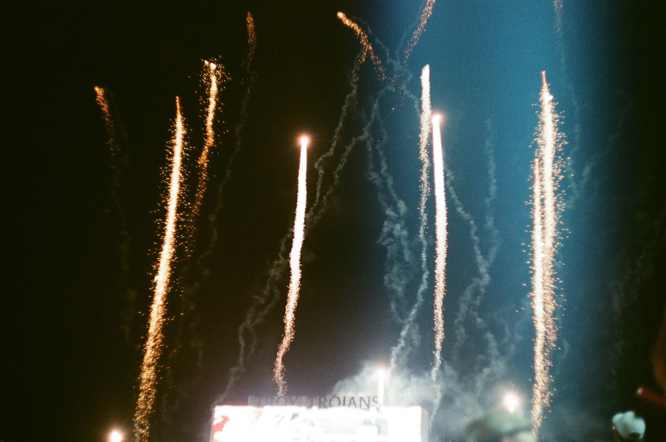 Night sky with fireworks leaving trails of sparks and smoke, with bright explosions and some faint billboards or signs visible at the bottom.