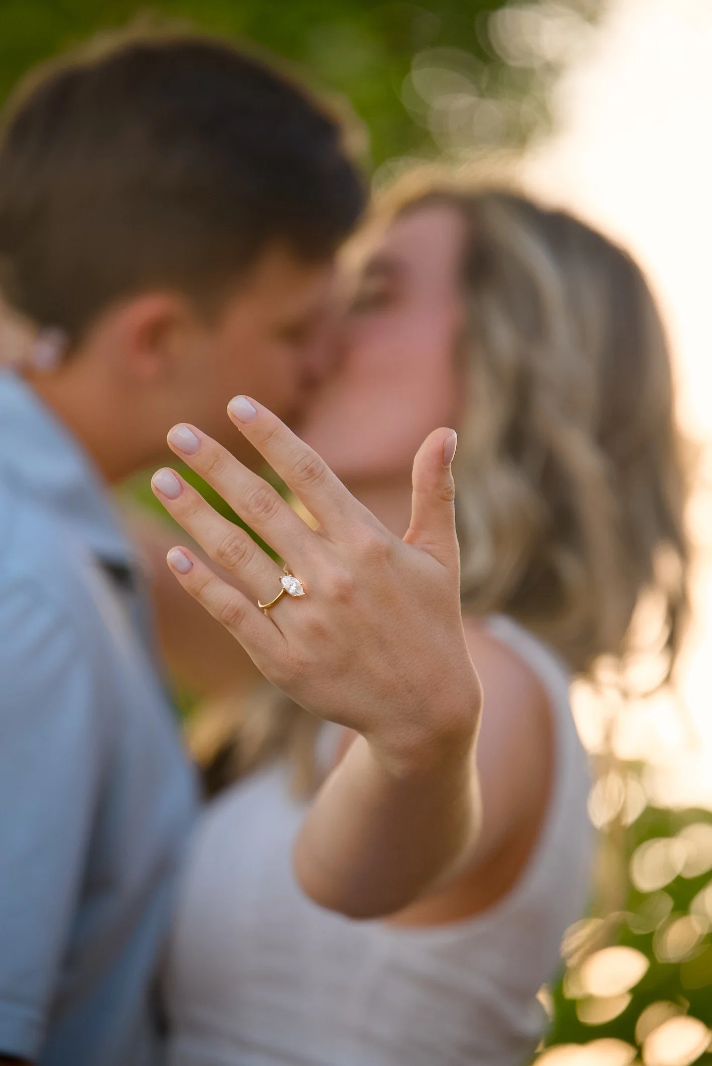 A woman holding hands with a man, showing an engagement ring on her finger, with a blurred couple in the background during the golden hour.