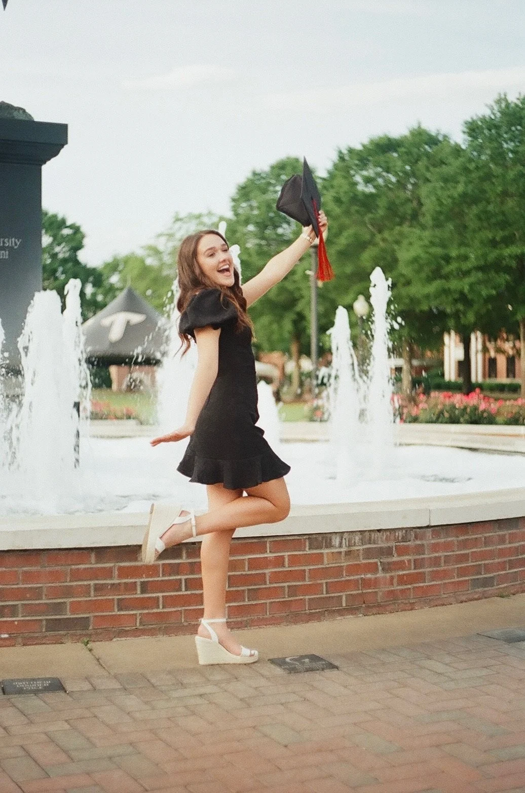 A young woman in a black dress with puffed sleeves and white wedge sandals is celebrating outdoors, jumping on one leg while holding a graduation cap in the air near a fountain with water jets and trees in the background.