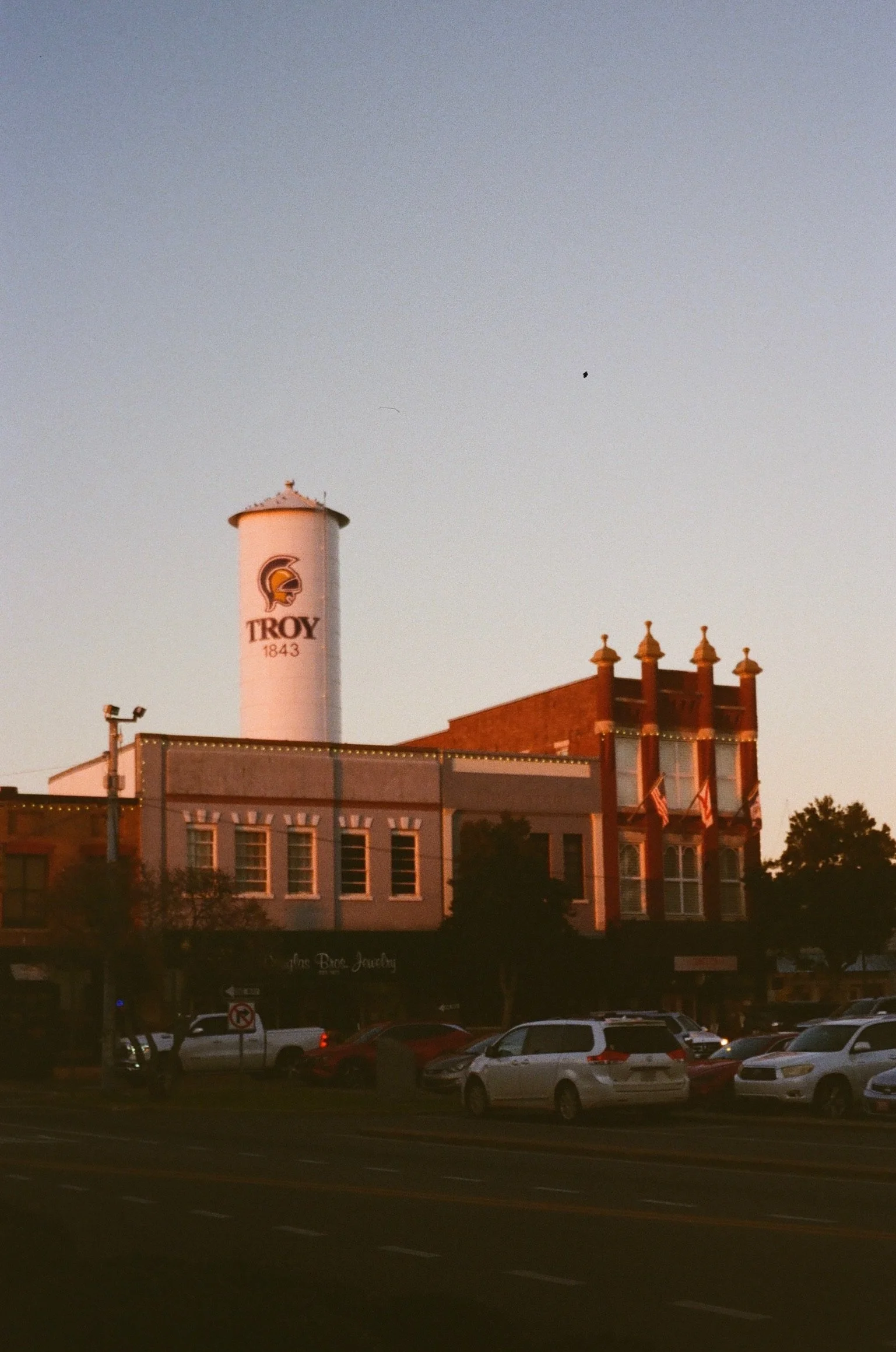 A street view of a historic building with a water tower that has a Troy University logo and the year 1843. The building has decorative brickwork and multiple chimneys, and there are several cars parked in the foreground.