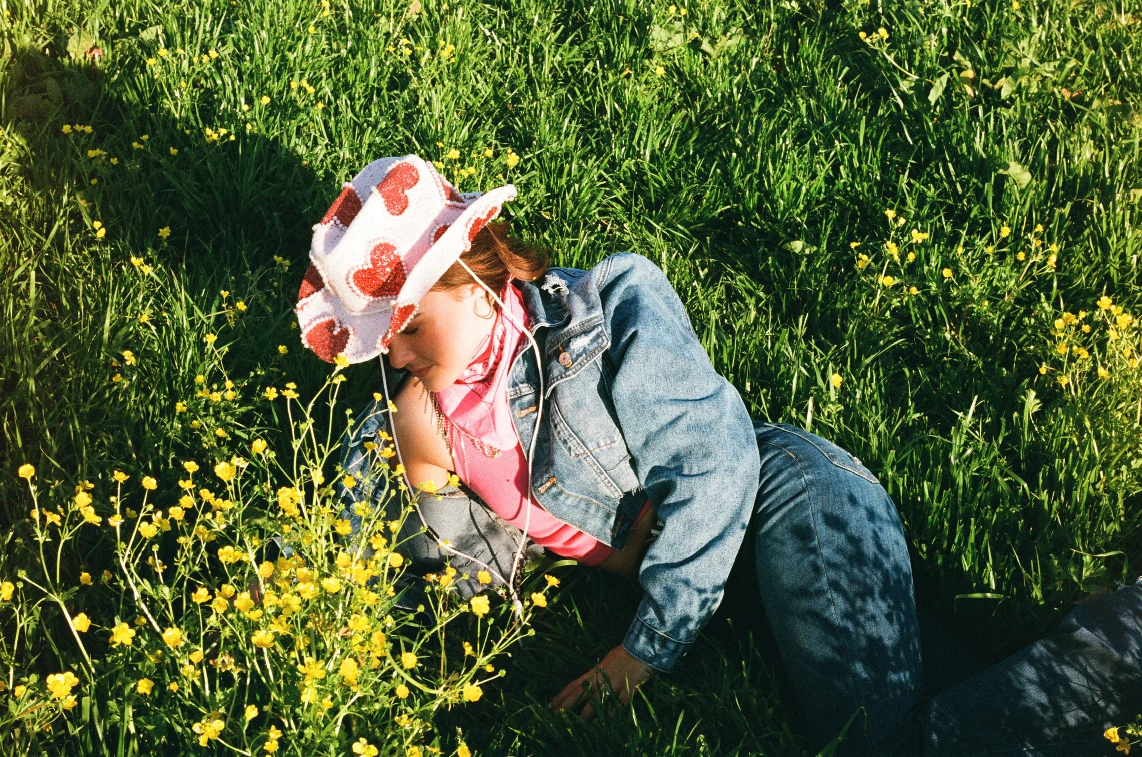 Child lying in a field of yellow flowers wearing a polka dot hat, pink shirt, denim jacket, and jeans, listening to music.