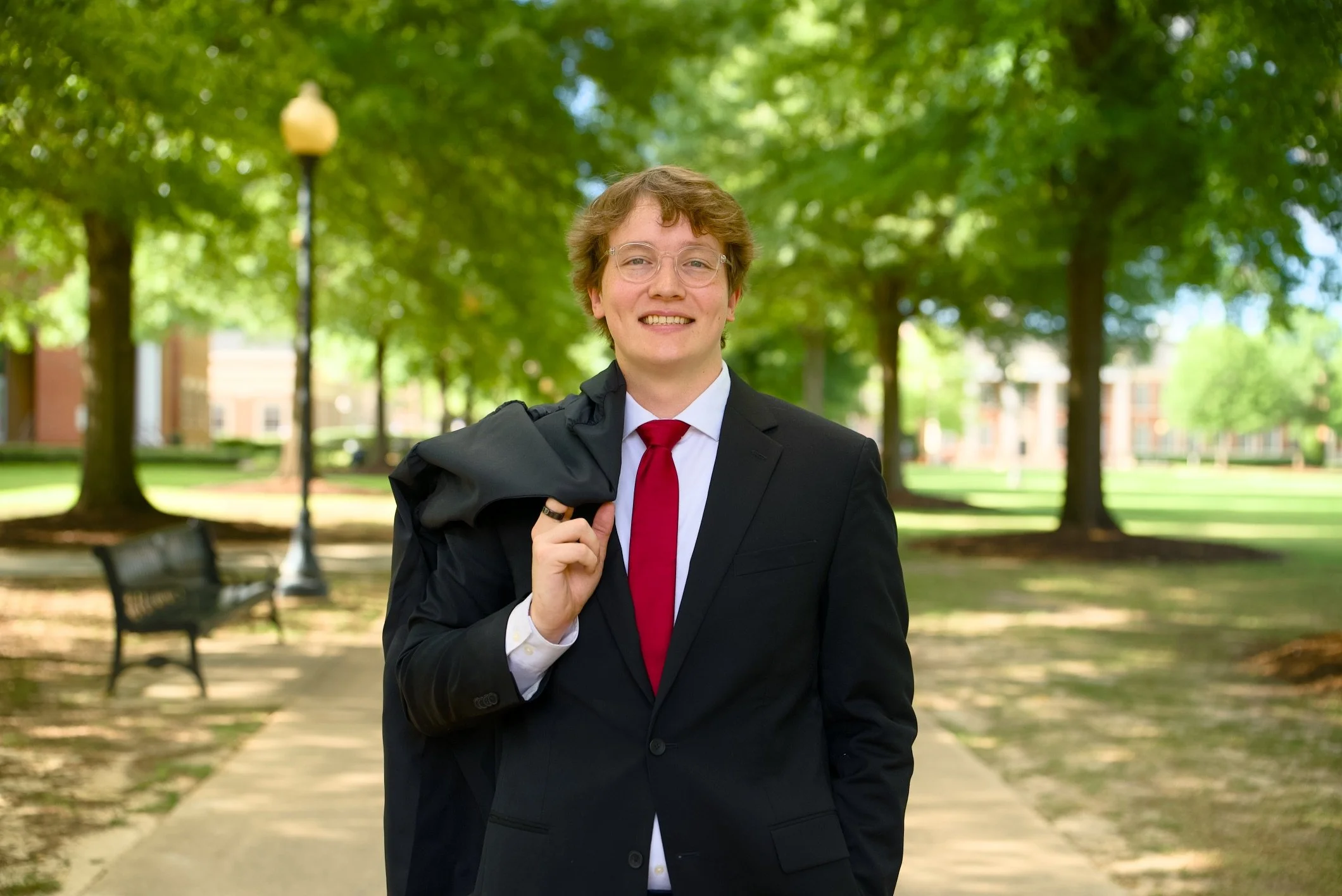 A young man in a business suit holding a jacket over his shoulder, smiling outdoors in a park with green trees and benches.