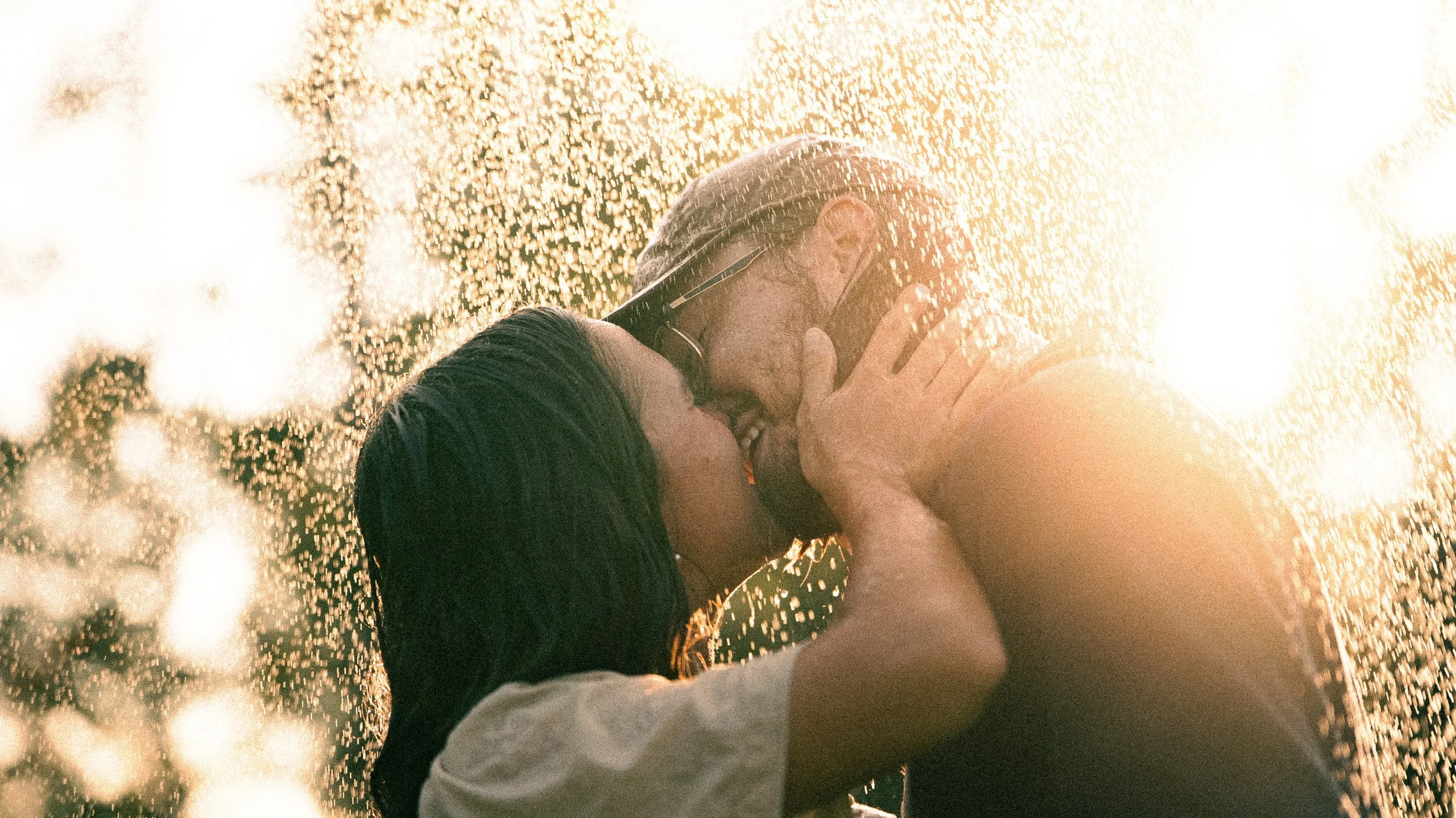 Two people kissing, one wearing a black shirt and glasses, the other with their hand on the other's face, with sunlight and water droplets creating a glowing effect.