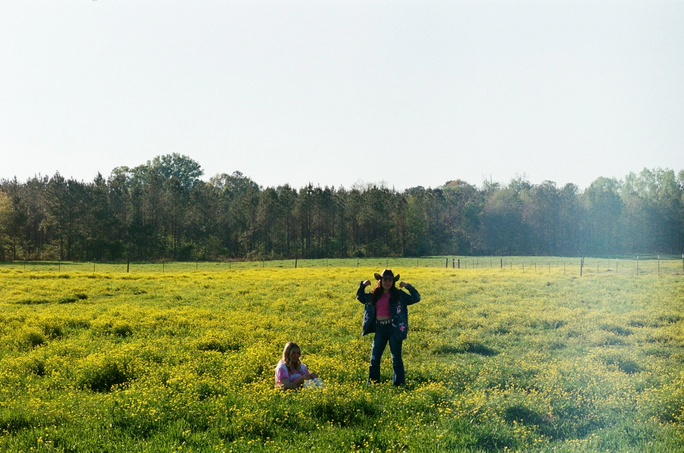 Two children in a field of yellow flowers, with a woman in the background. The woman is wearing a cowboy hat and a jacket. In the distance, there are trees and a fence under a clear sky.