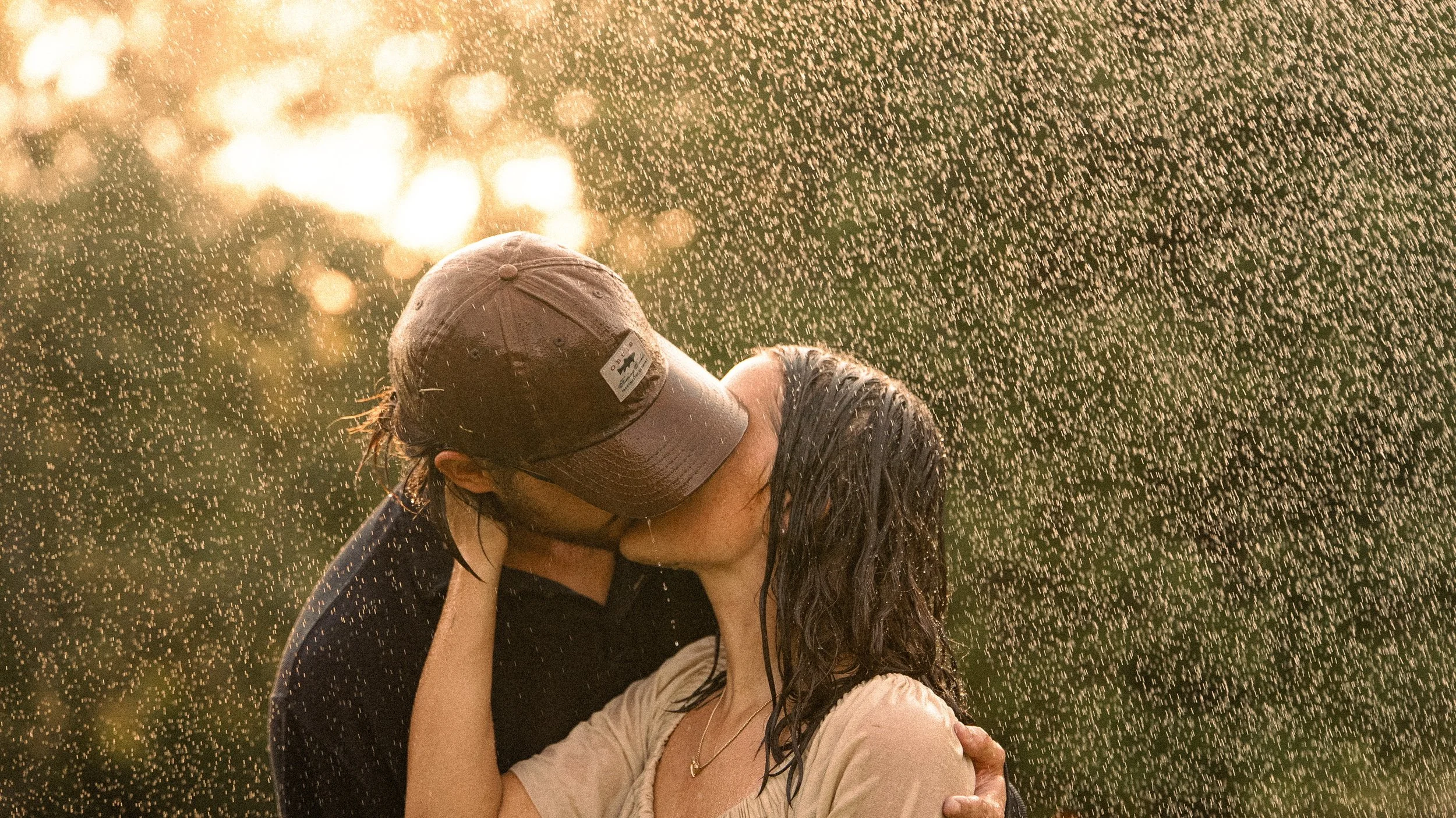A couple embracing in the rain with sunlight shining through, the woman wearing a beige top and the man wearing a cap and dark clothing, both wet from the rain.