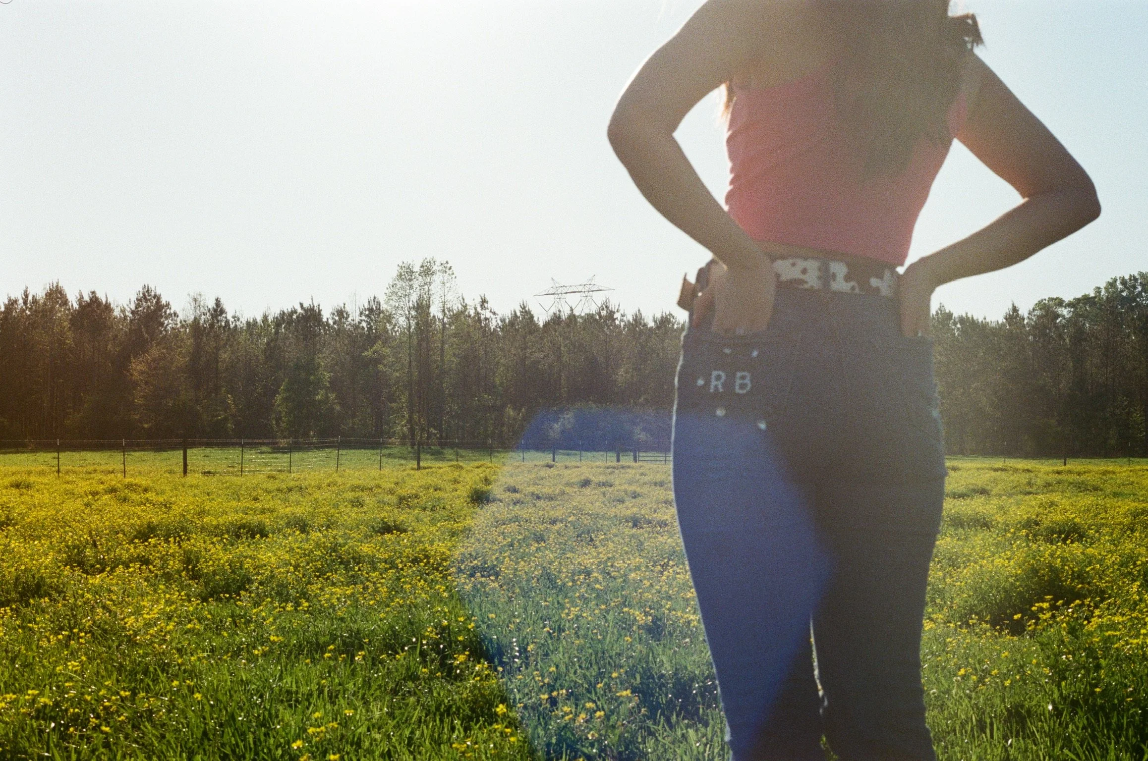 A person standing in a field of yellow flowers with trees and a power line in the background. The person has long curly hair and is wearing a pink top, patterned underwear, and black pants with white text.