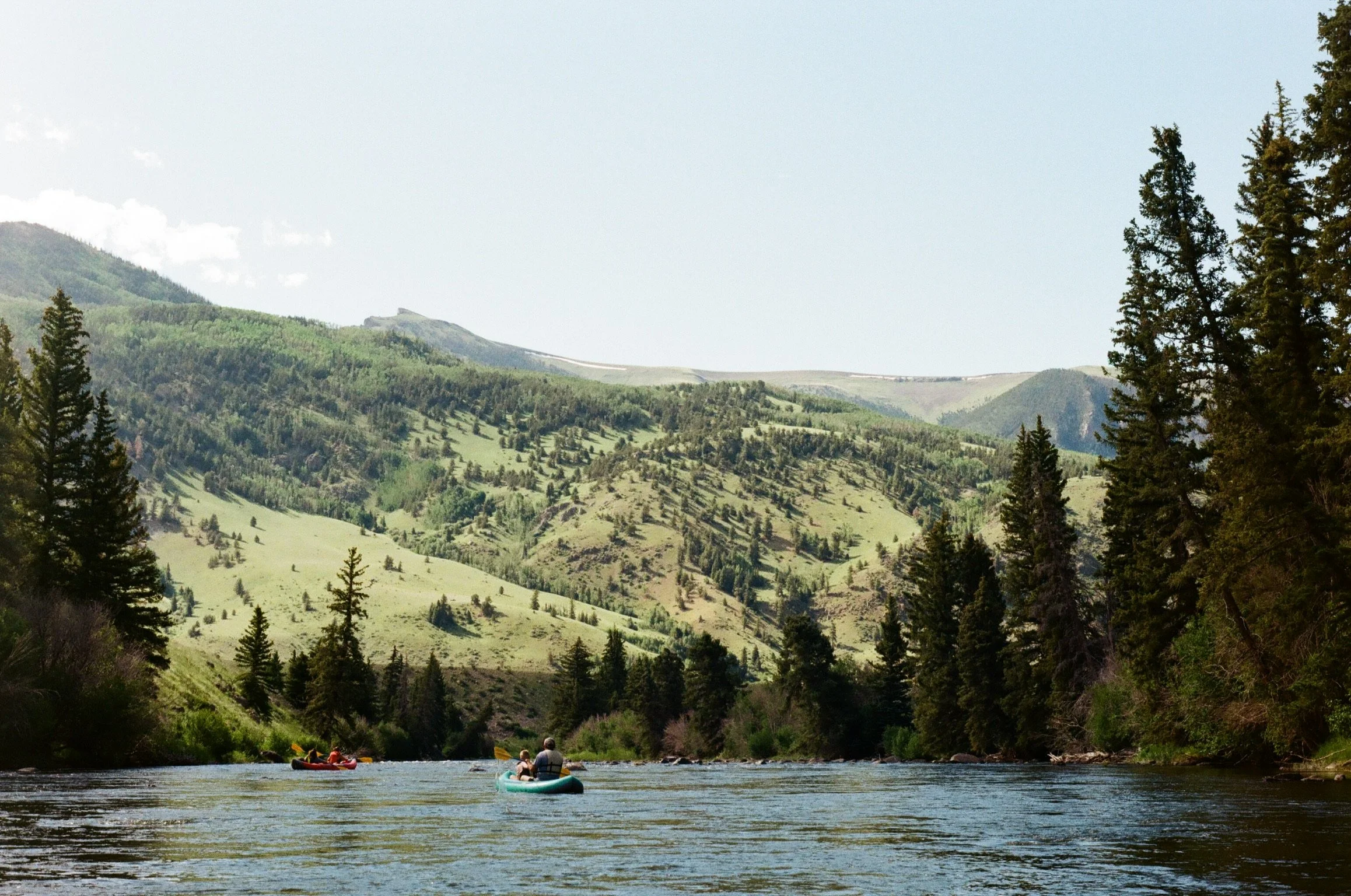 People kayaking on a river surrounded by lush green hills and tall pine trees, with mountains in the background under a partly cloudy sky.