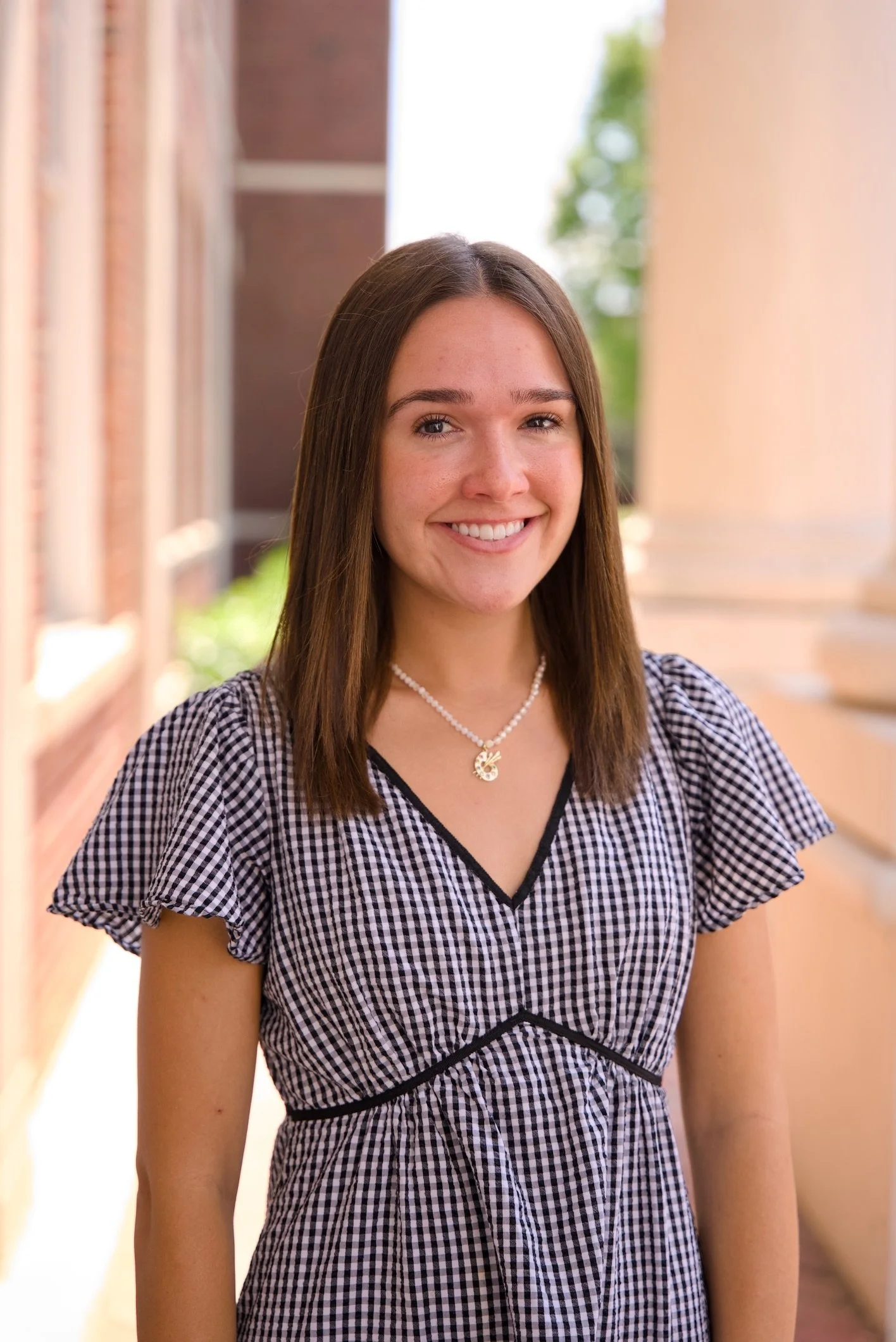Portrait of a young woman with long brown hair smiling outside, wearing a black and white checkered dress and a pearl necklace, with columns and greenery in the background.
