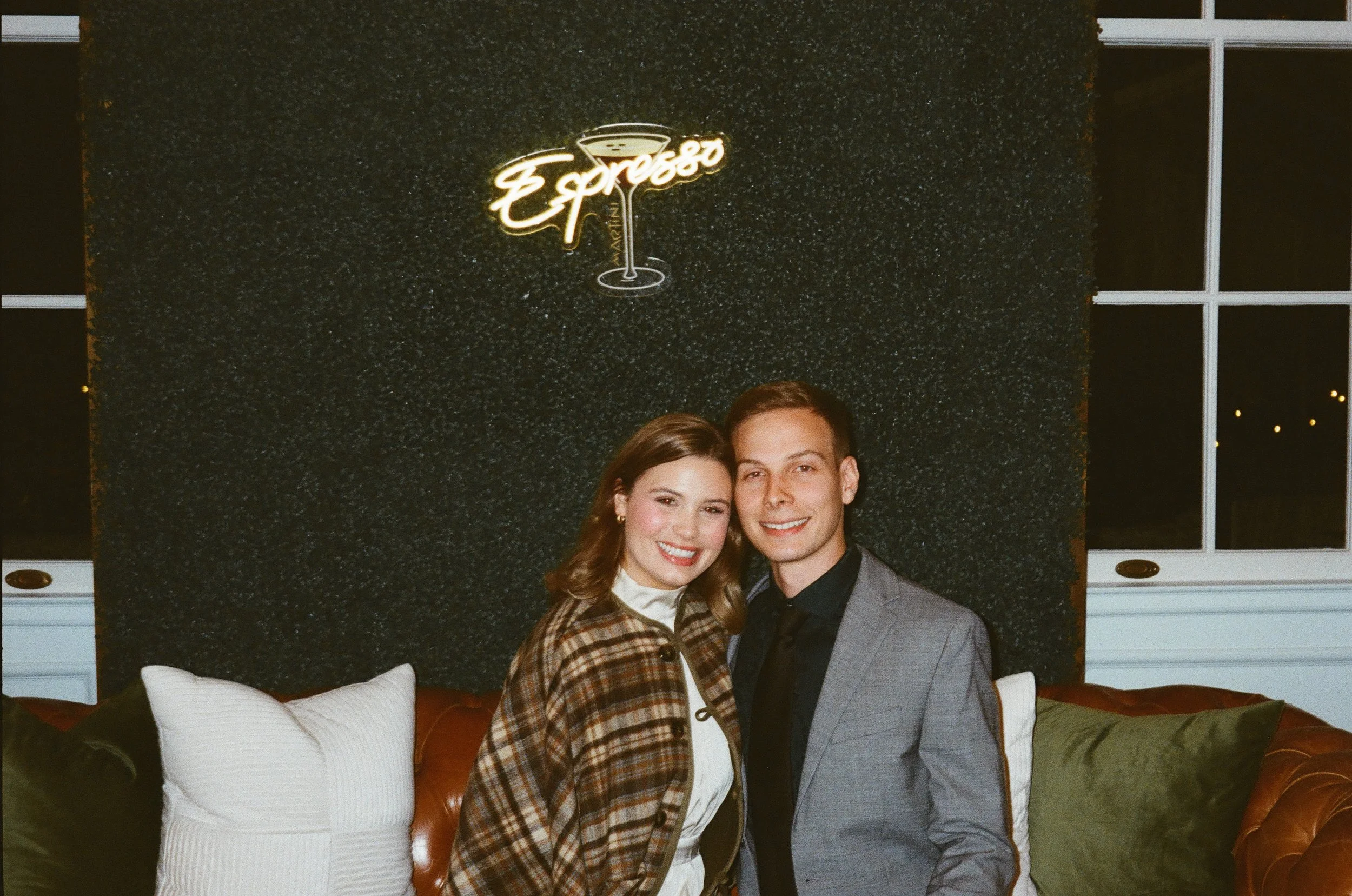 A young couple standing close together in a cozy indoor setting, smiling at the camera. The woman has shoulder-length brown hair and is wearing a plaid jacket over a white top. The man has short dark hair and is dressed in a gray suit with a black sh