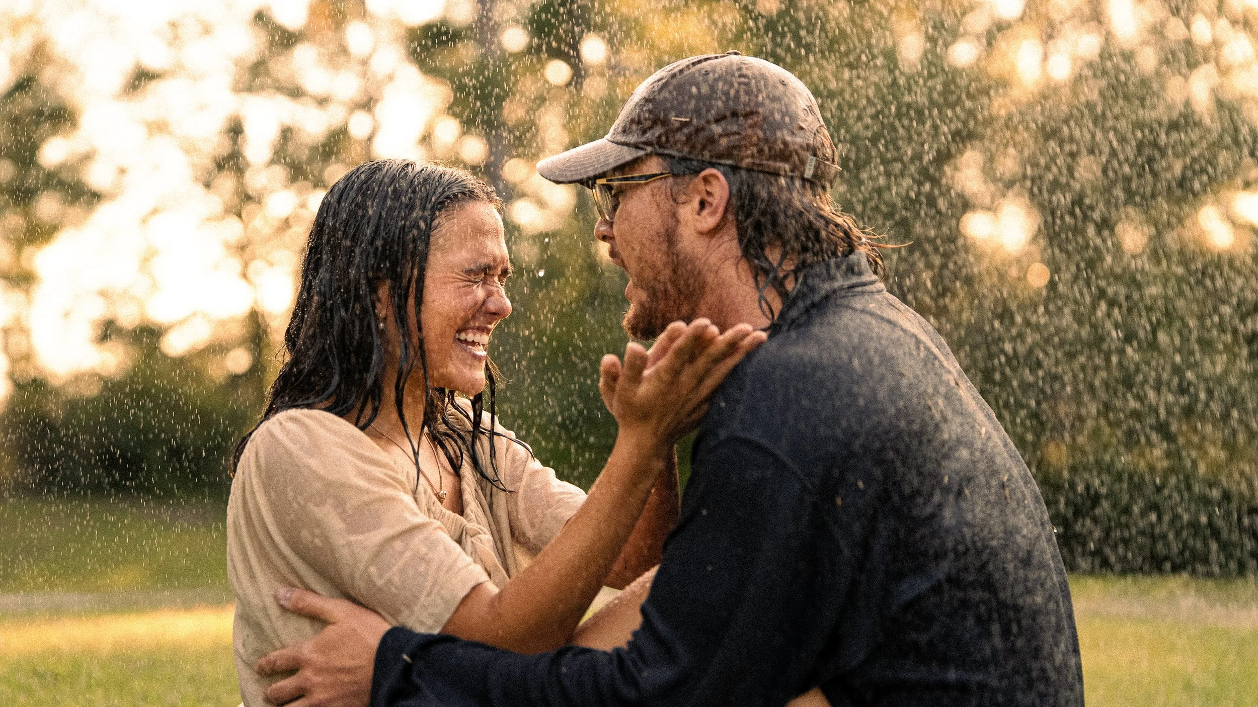 A man and woman smiling and leaning into each other in the rain, with trees and sunlight in the background.