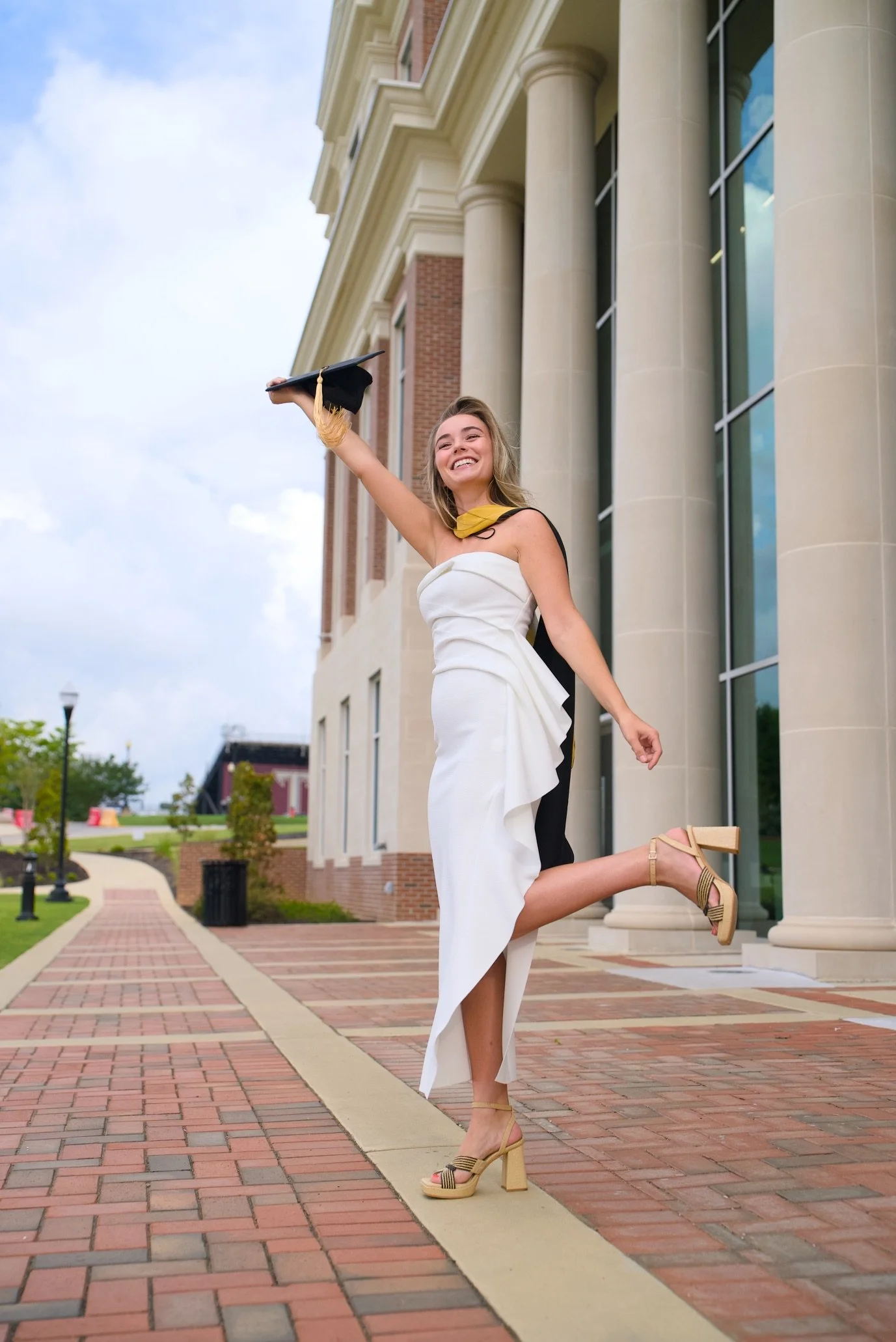 A young woman with blonde hair, dressed in a white dress with black accents, is celebrating graduation outside a large, classical-style building with tall columns and brick walls. She is holding a graduation cap in her raised hand and has a joyful ex