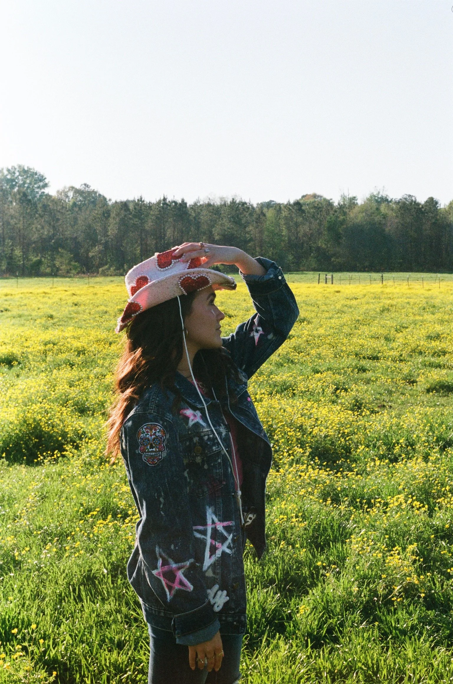 A woman wearing a colorful hat with red hearts and white flowers, a denim jacket with patches, standing in a field of yellow flowers with trees in the background, holding her hand to her hat.