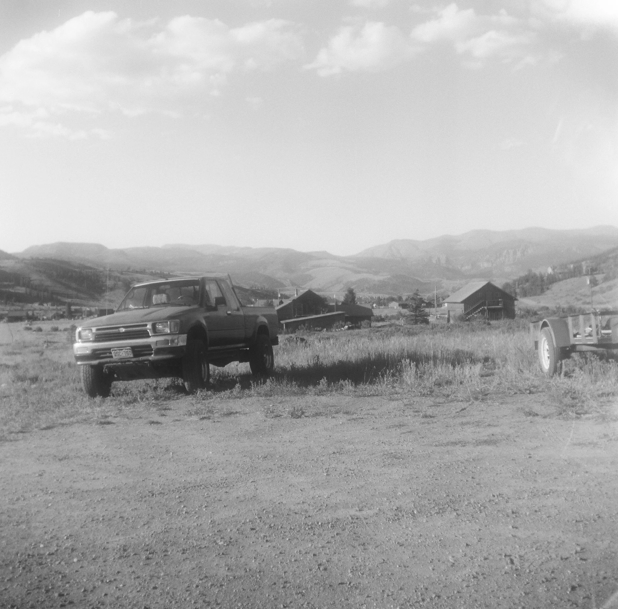 Black and white photo of a rural area with mountains in the background, a car, houses, and a gravel open lot.