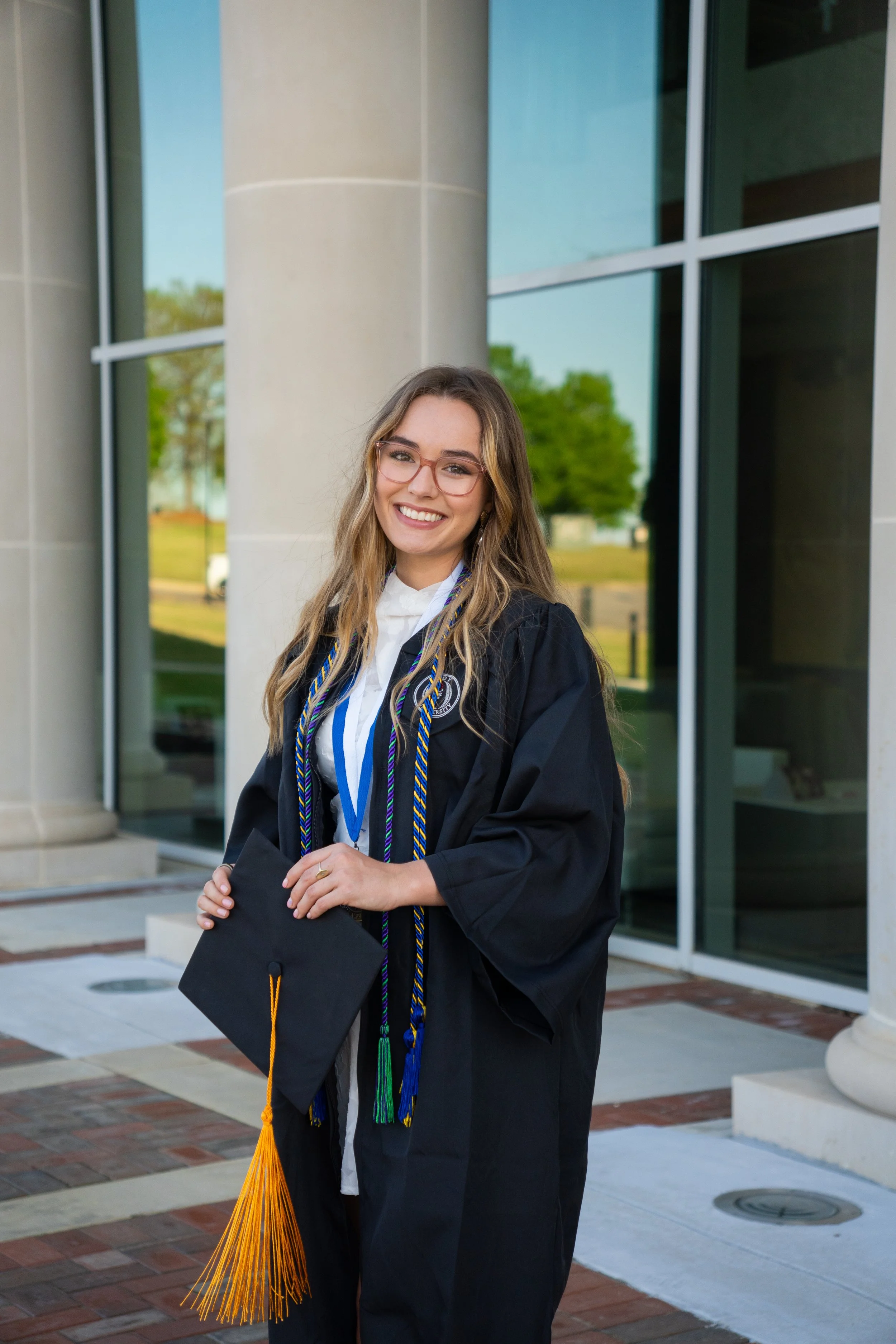 A young woman in graduation cap and gown standing outside a building with large glass windows, smiling while holding her diploma, with graduation cords around her neck.
