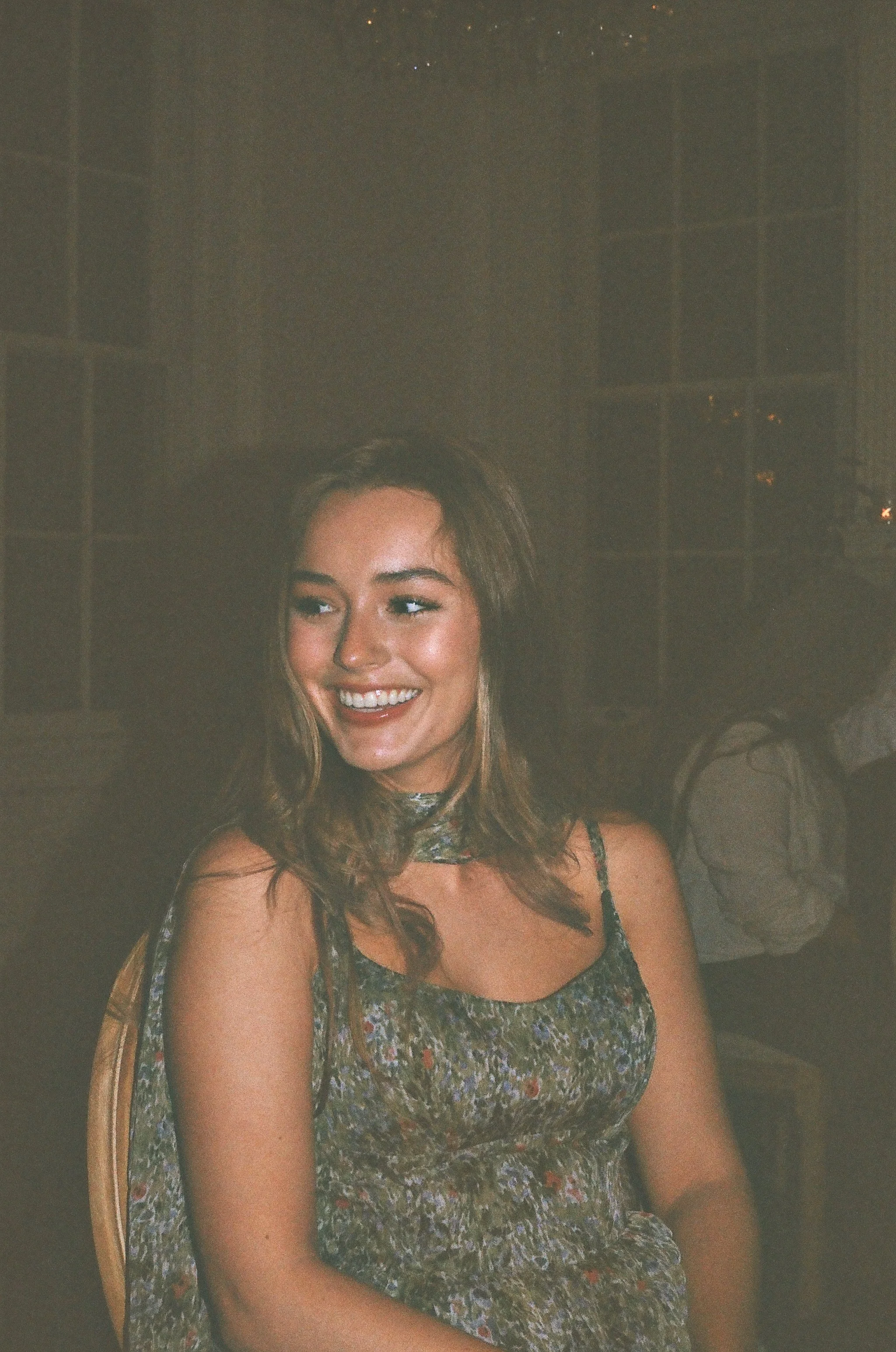 A woman with long wavy brown hair smiling, wearing a colorful sleeveless dress, sitting in a room with a dark grid-patterned wall in the background.