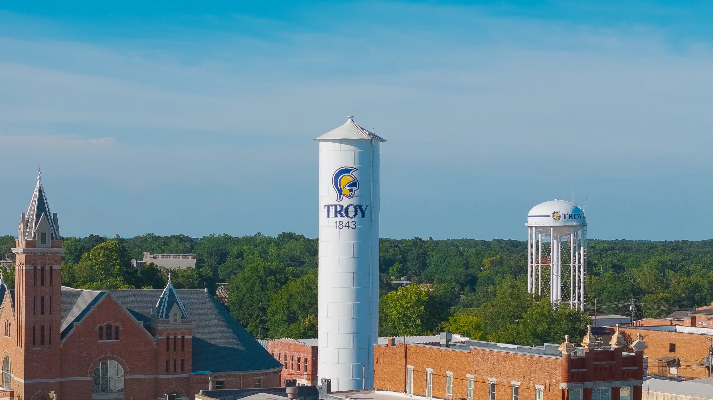 A water tower and a cylindrical tank, both featuring the Troy University logo, with a church and green trees in the background under a blue sky.