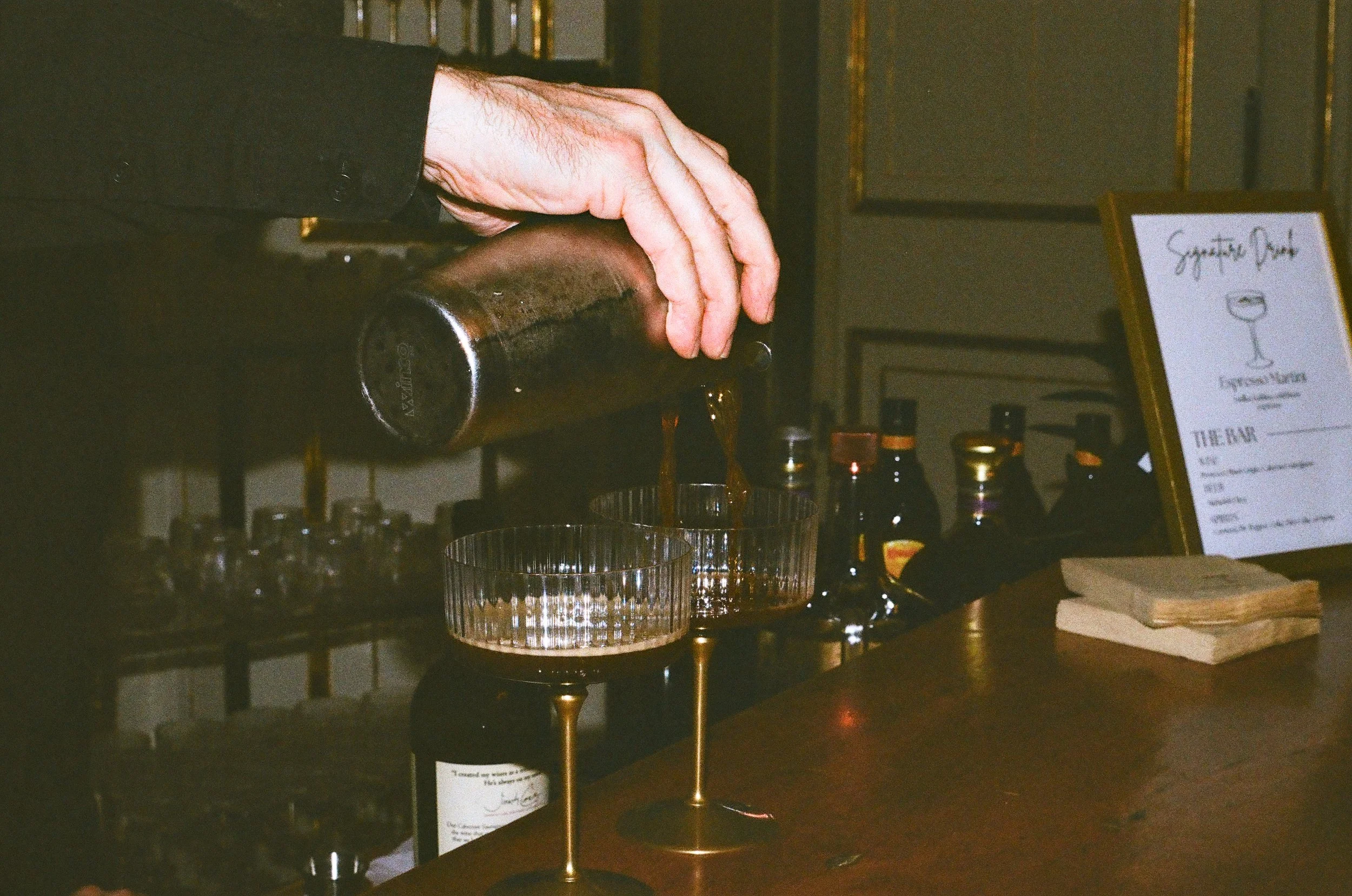 A hand pouring a dark beverage into a glass on a bar counter, with a sign and various bottles in the background.