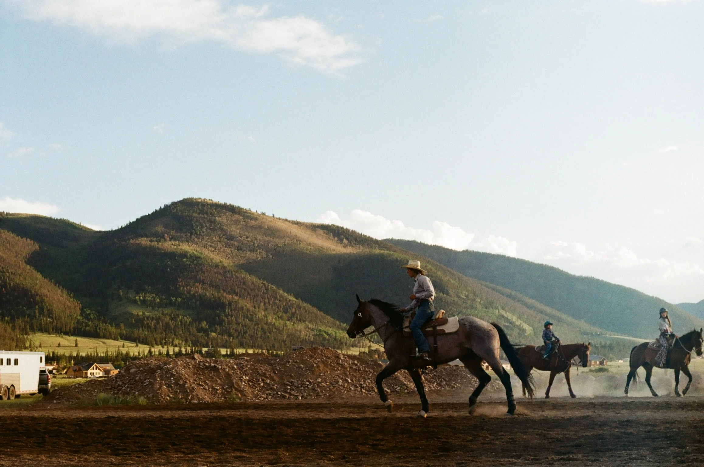 People riding horses on a dirt track with mountains and houses in the background under a partly cloudy sky.