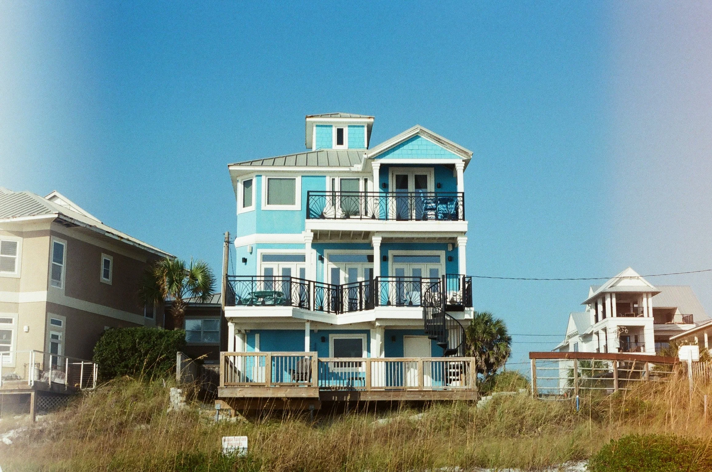 A tall, blue beach house with multiple balconies and white trim, situated near the beach under a clear blue sky.