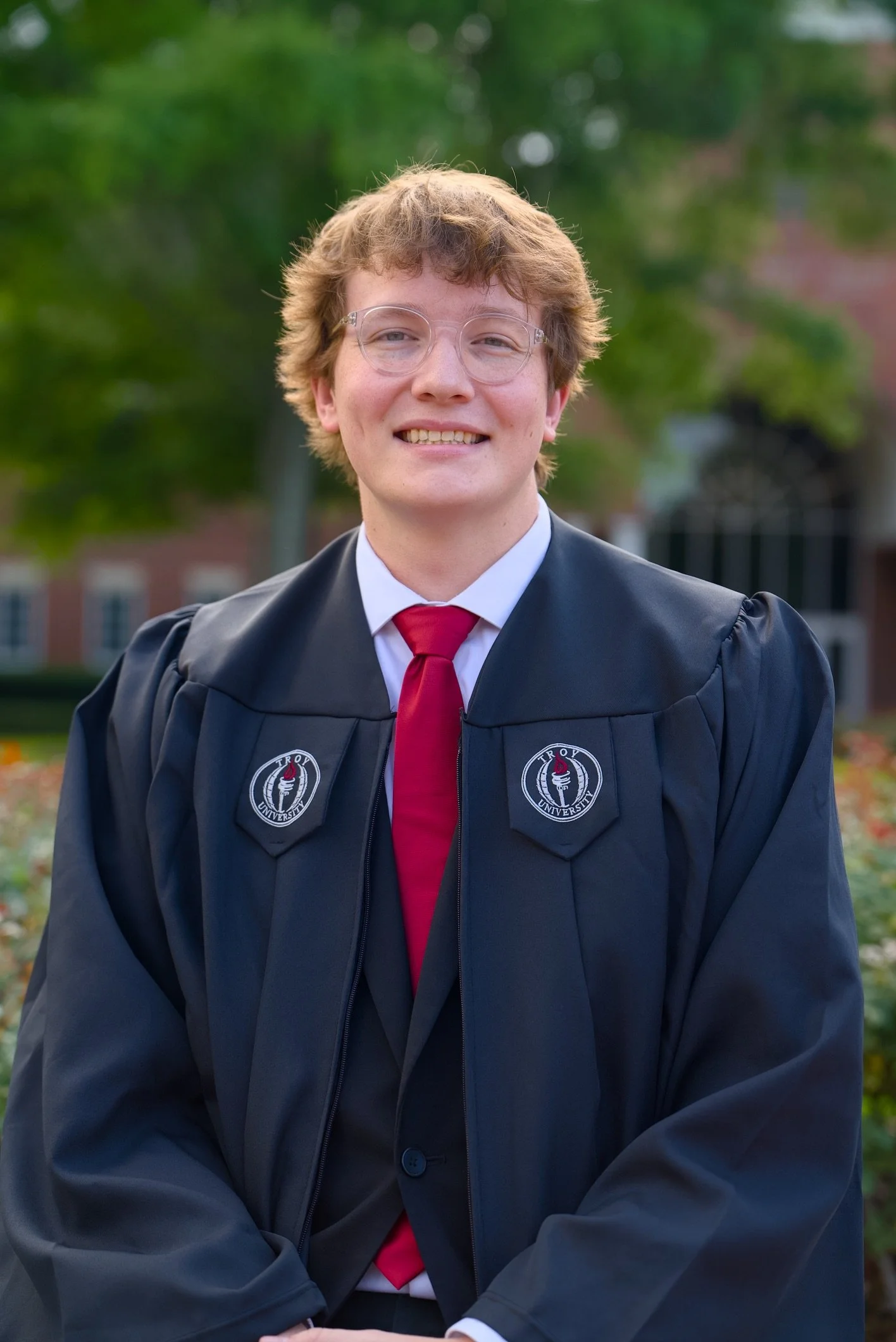 A young man wearing glasses, a black graduation gown with a 'Troy University' emblem, white shirt, and red tie, smiling outdoors with blurred green trees in the background.