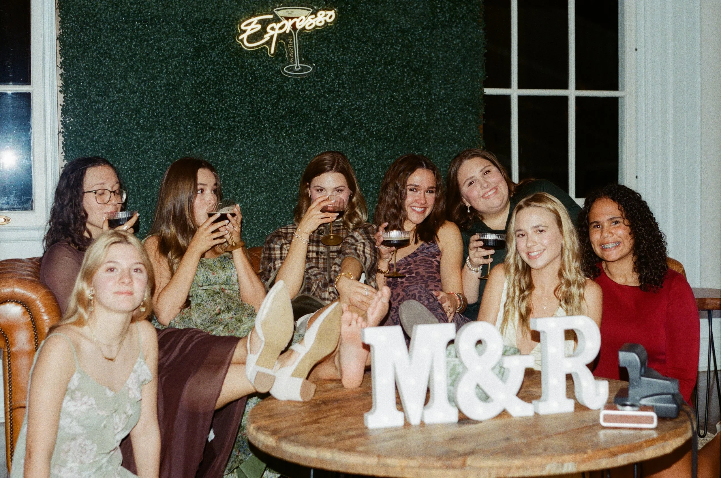 A group of young women sitting together at a party, holding drinks and smiling for the camera. The background features a green artificial hedge wall with a neon sign that reads 'Espresso' and a cocktail glass graphic.