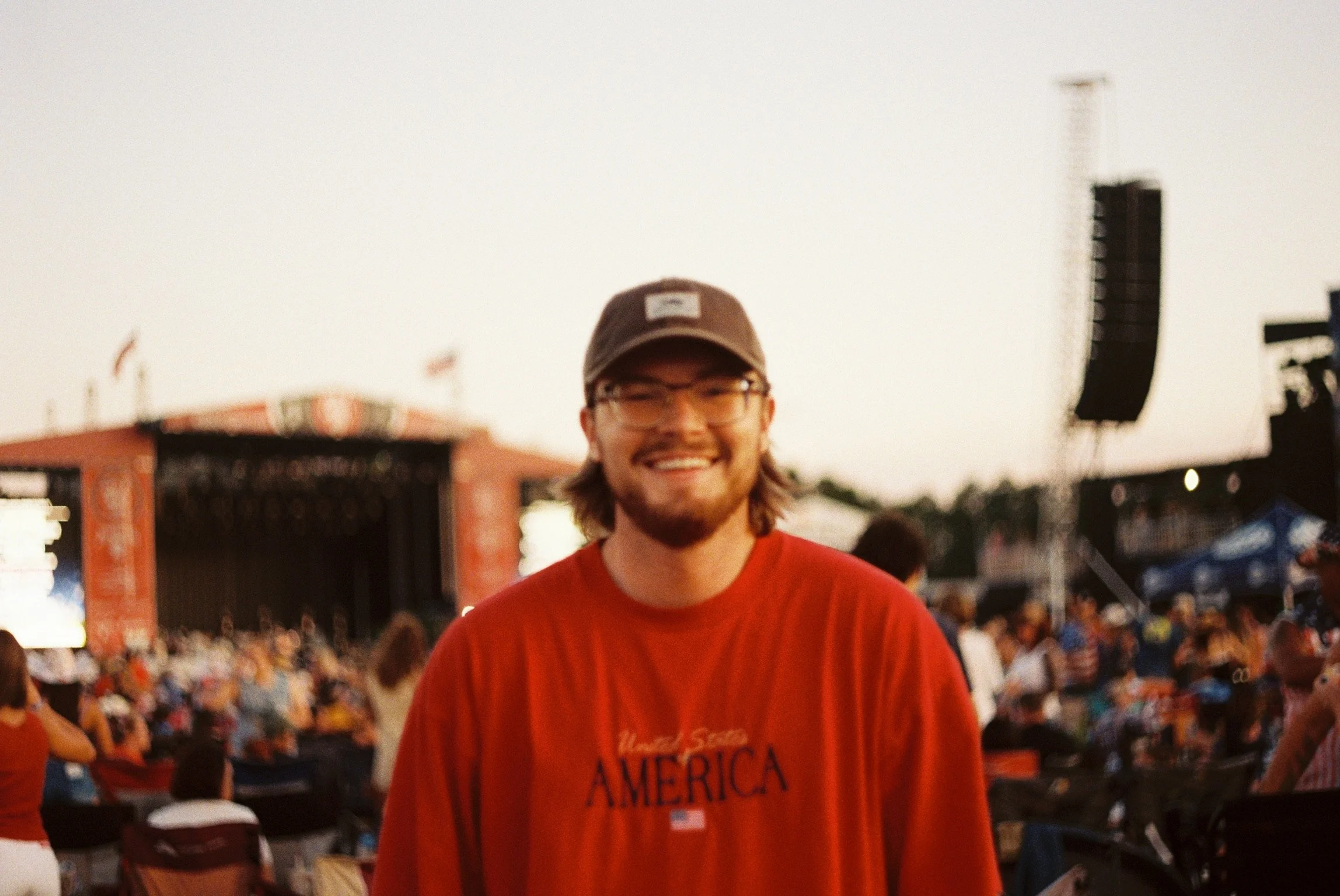 A smiling man with long hair, glasses, and a cap, wearing a red shirt that says 'America,' at an outdoor event with a crowd and stage in the background during the evening.