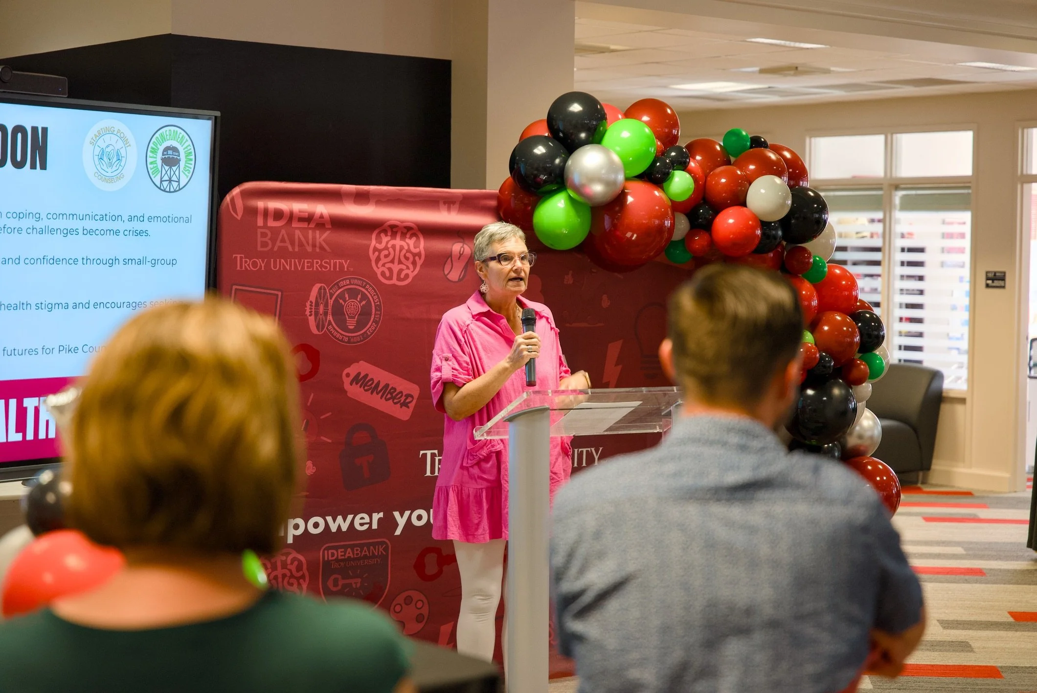 A woman in a pink outfit speaking into a microphone at an indoor event, with a balloon arch and a large screen displaying presentation slides in the background. The audience is seated and facing her.