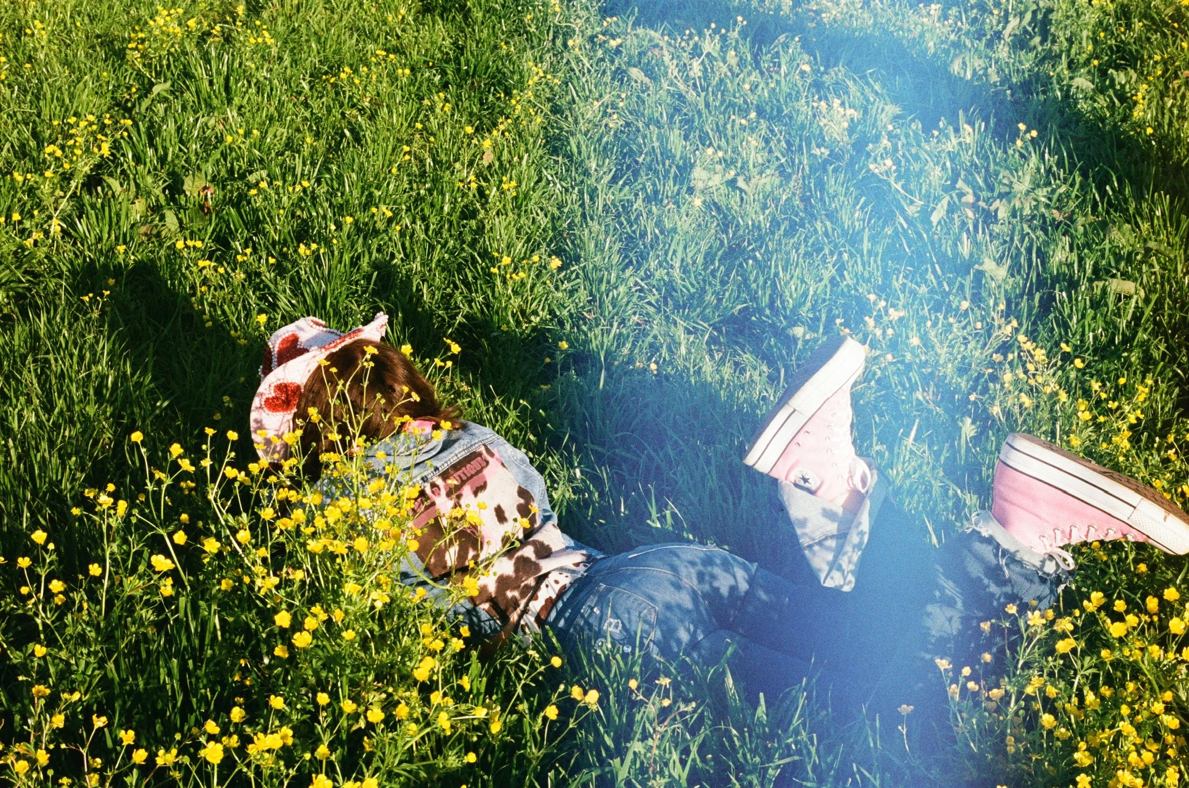 Person lying in a field of yellow wildflowers with white sneakers visible, wearing jeans and a patterned scarf, basking in sunlight.