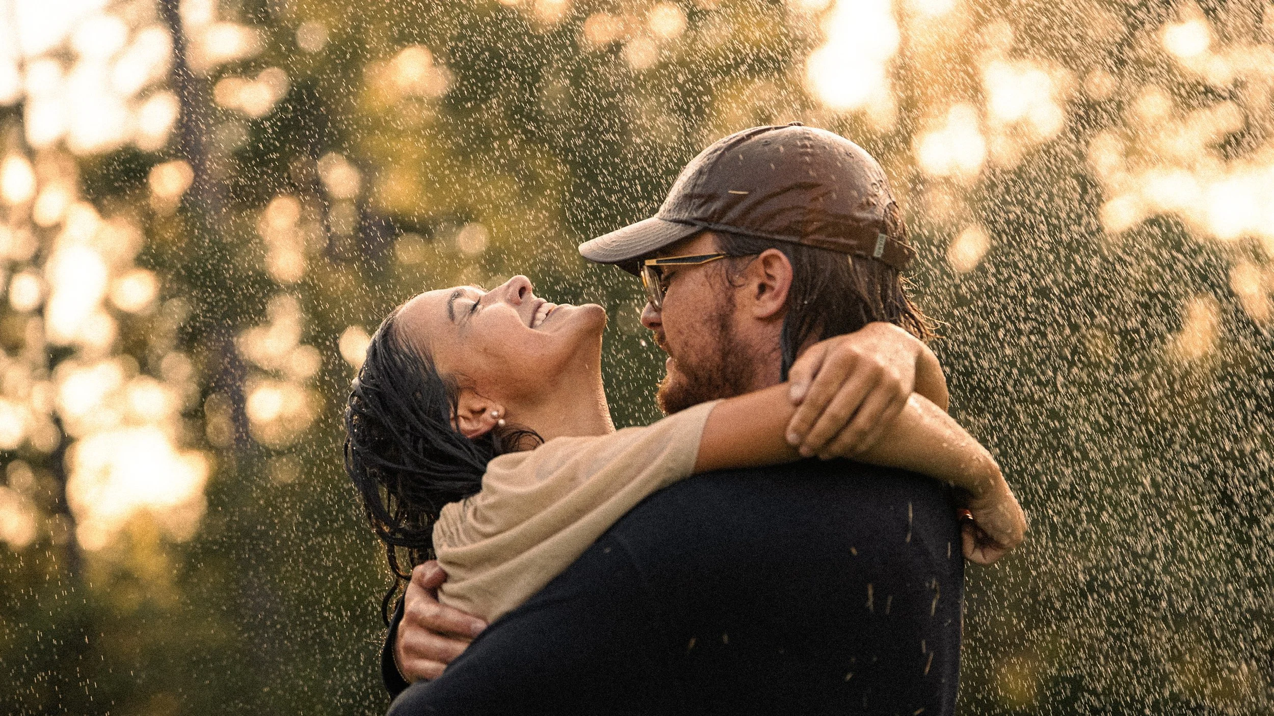 A man and woman smiling and embracing in the rain at sunset, with water droplets visible and blurred golden trees in the background.