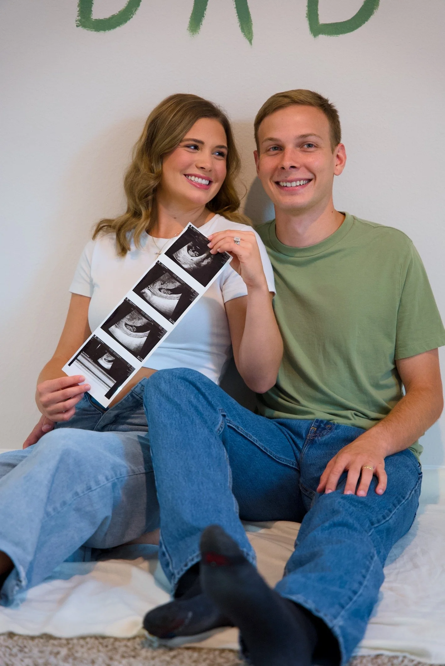 A smiling couple sitting on a bed, holding ultrasound pictures, with a wall background and partially visible green text above them.