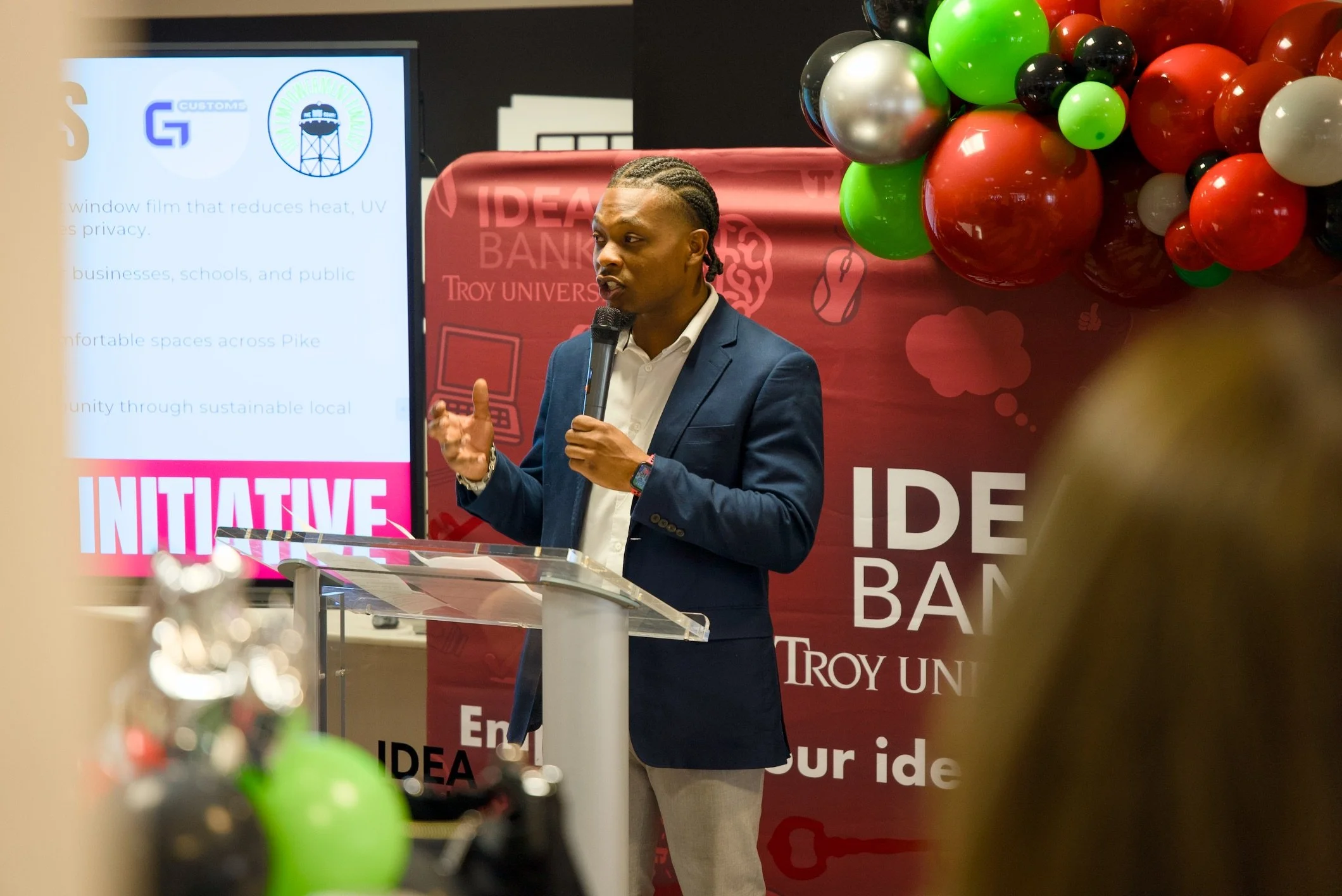 A man in a suit and white shirt speaking into a microphone at a presentation. There are colorful balloons and a red banner behind him with partially visible text, and a large display screen with logos and text.