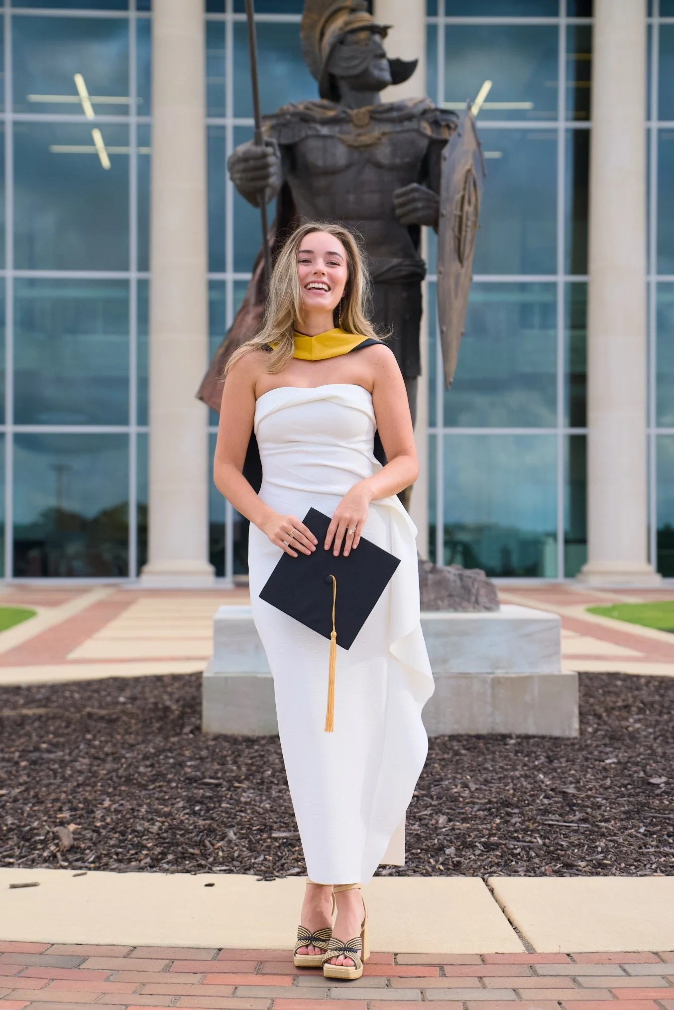 A young woman in a white dress with a yellow neck scarf, holding a diploma, standing in front of a large statue of a warrior. The woman is smiling and wearing platform sandals. The background features a modern building with large glass windows.