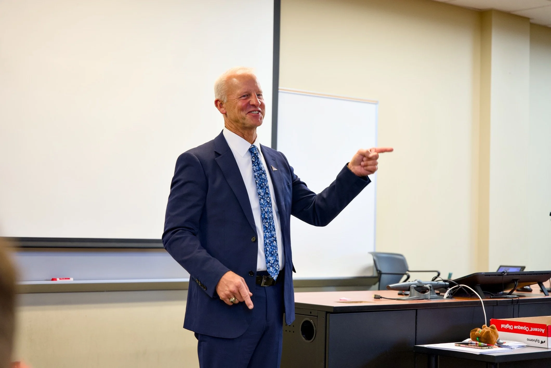 A man in a navy suit and tie is standing in front of a whiteboard in a classroom or conference room, smiling and pointing to the side.